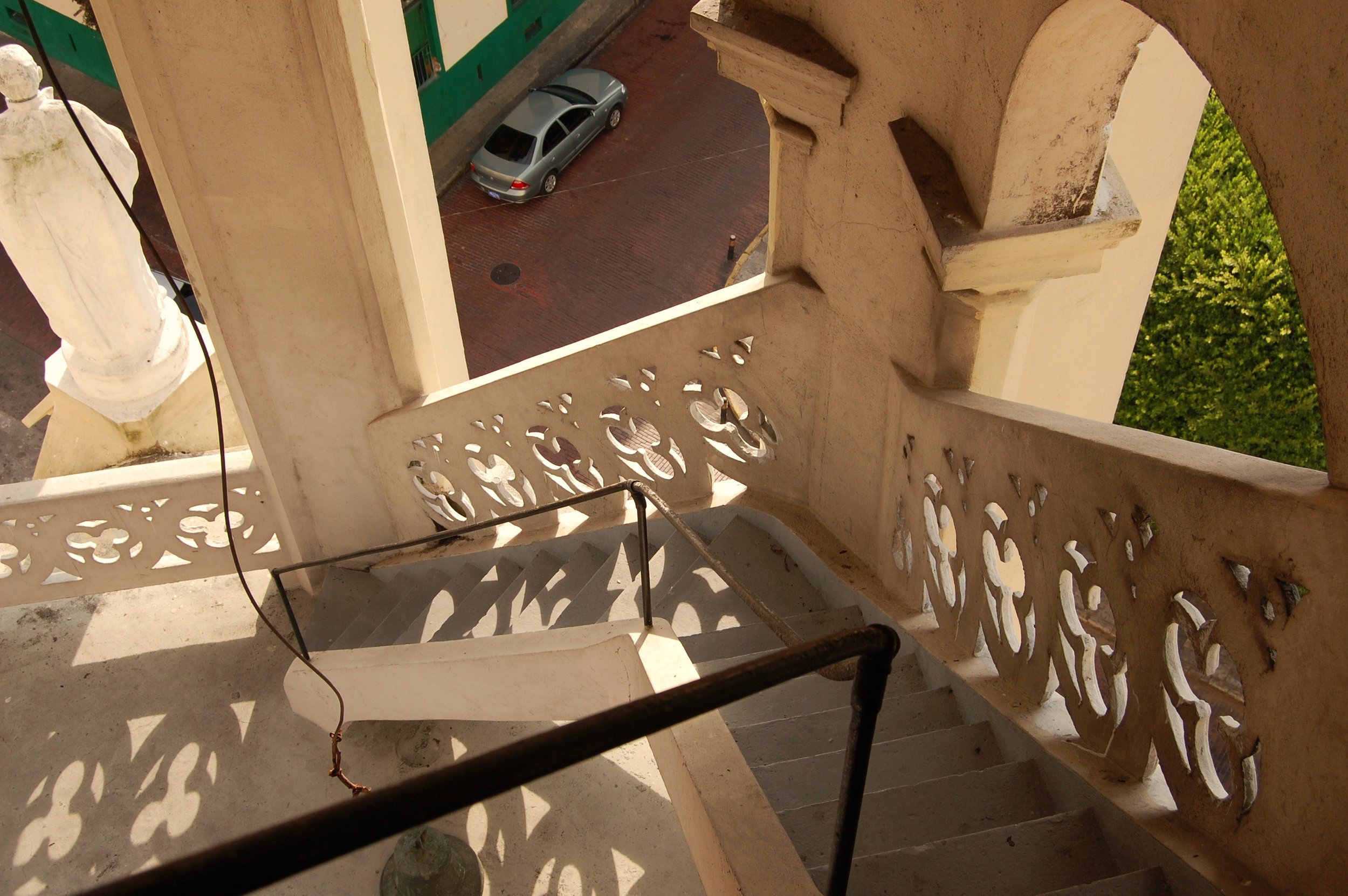 An upward view from a staircase looking down at a street with a parked silver car, part of a building, green door, some trees, and shadows cast by the staircase railing.