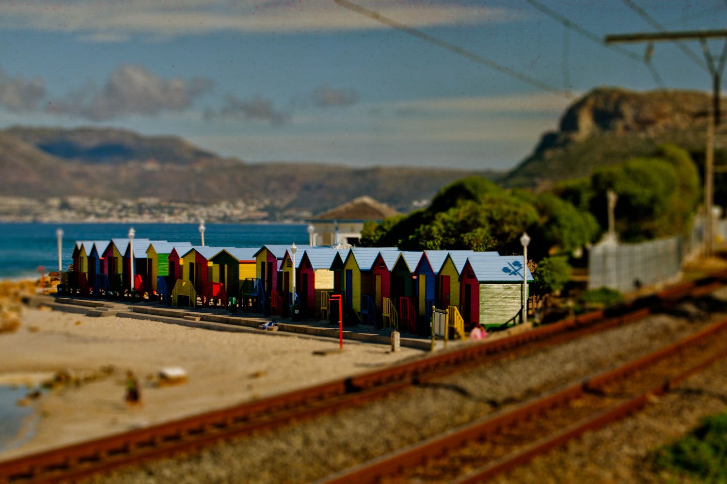 Colorful beach huts along a boardwalk with a train track in the foreground, green shrubbery, hills, and water in the background.