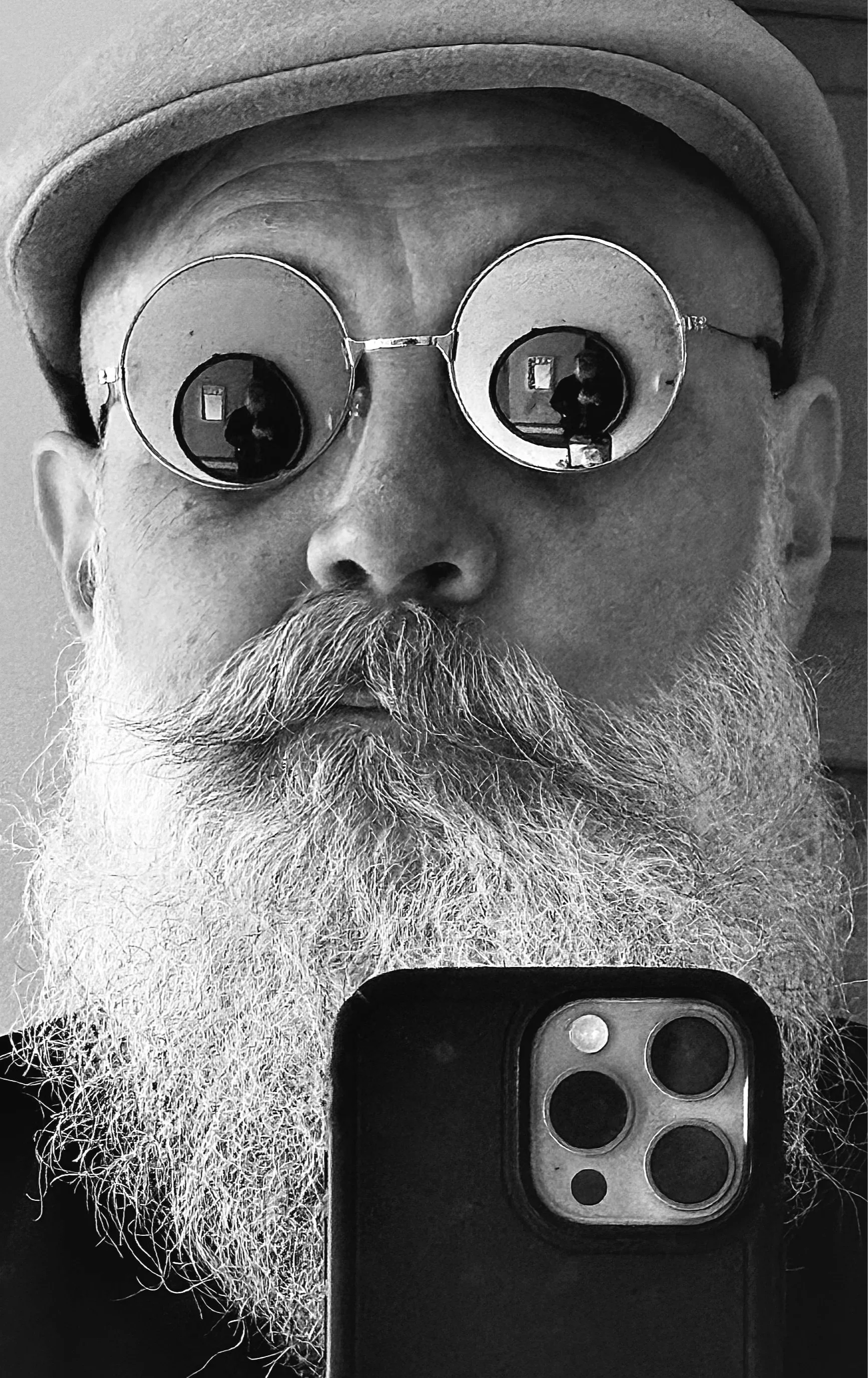 A black and white selfie of an older man with a beard, wearing glasses and a hat, holding a smartphone in front of his face. The reflection in his glasses shows the photographer taking the picture.