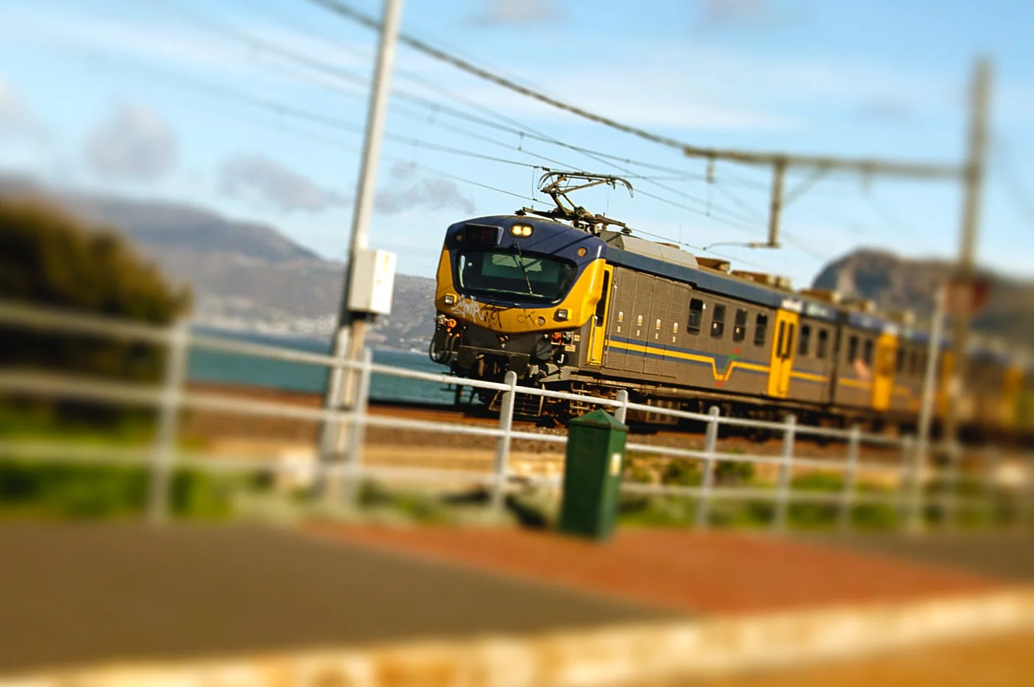 A yellow and black train traveling on a railway track near a body of water with mountains in the background.