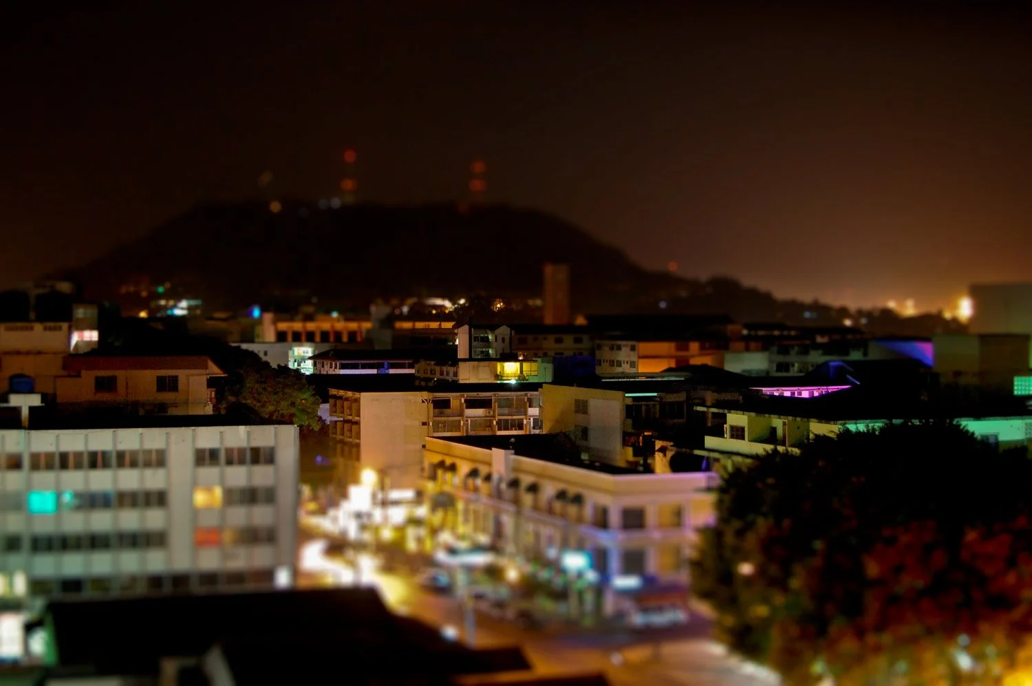 Nighttime cityscape with illuminated buildings and a mountain in the background with red lights on top.