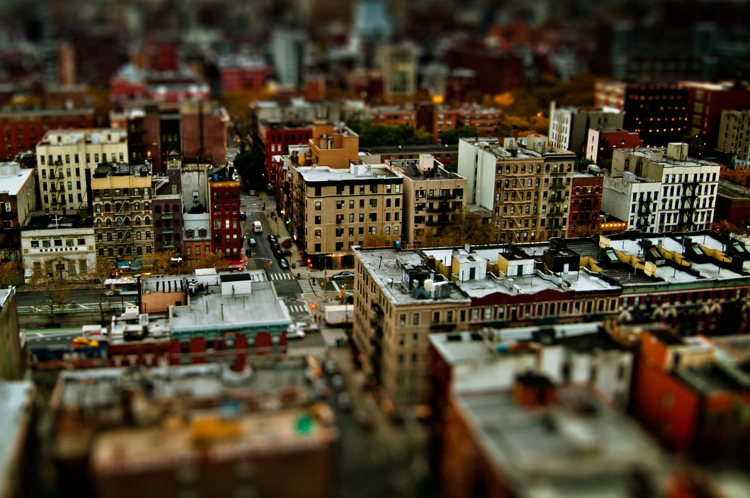 Aerial view of a cityscape with numerous tightly packed buildings, streets, and some trees, taken during the evening or early night with city lights starting to turn on.