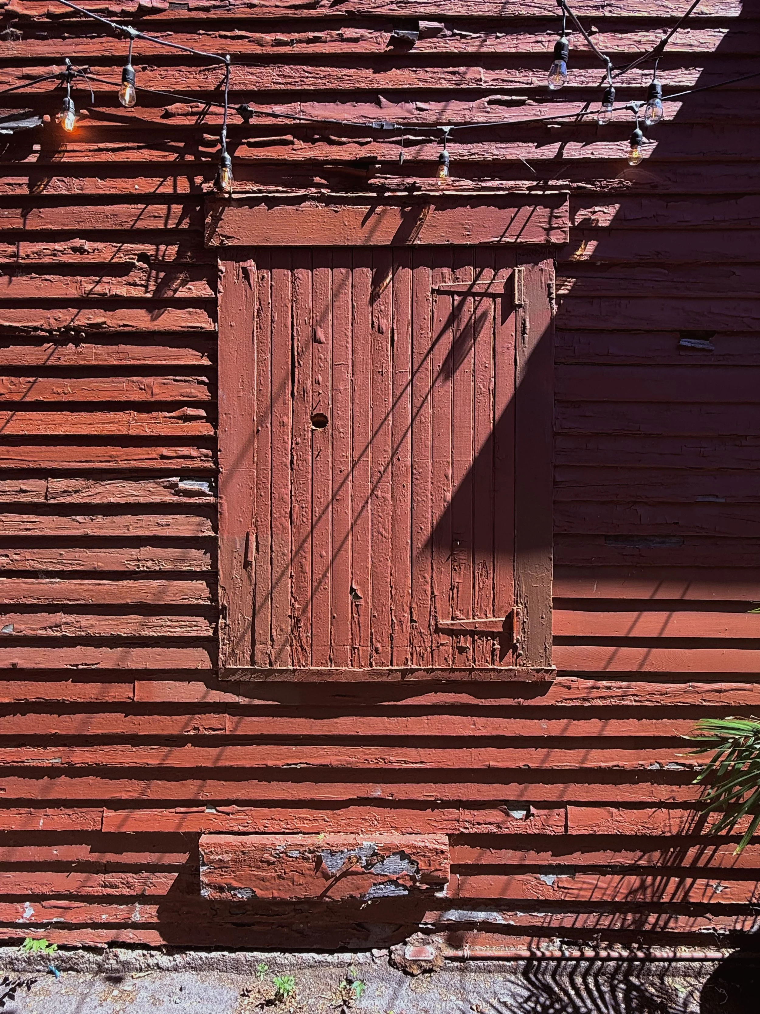 Old red wooden door with peeling paint on a red wooden wall, with shadows from string lights cast on the wall.