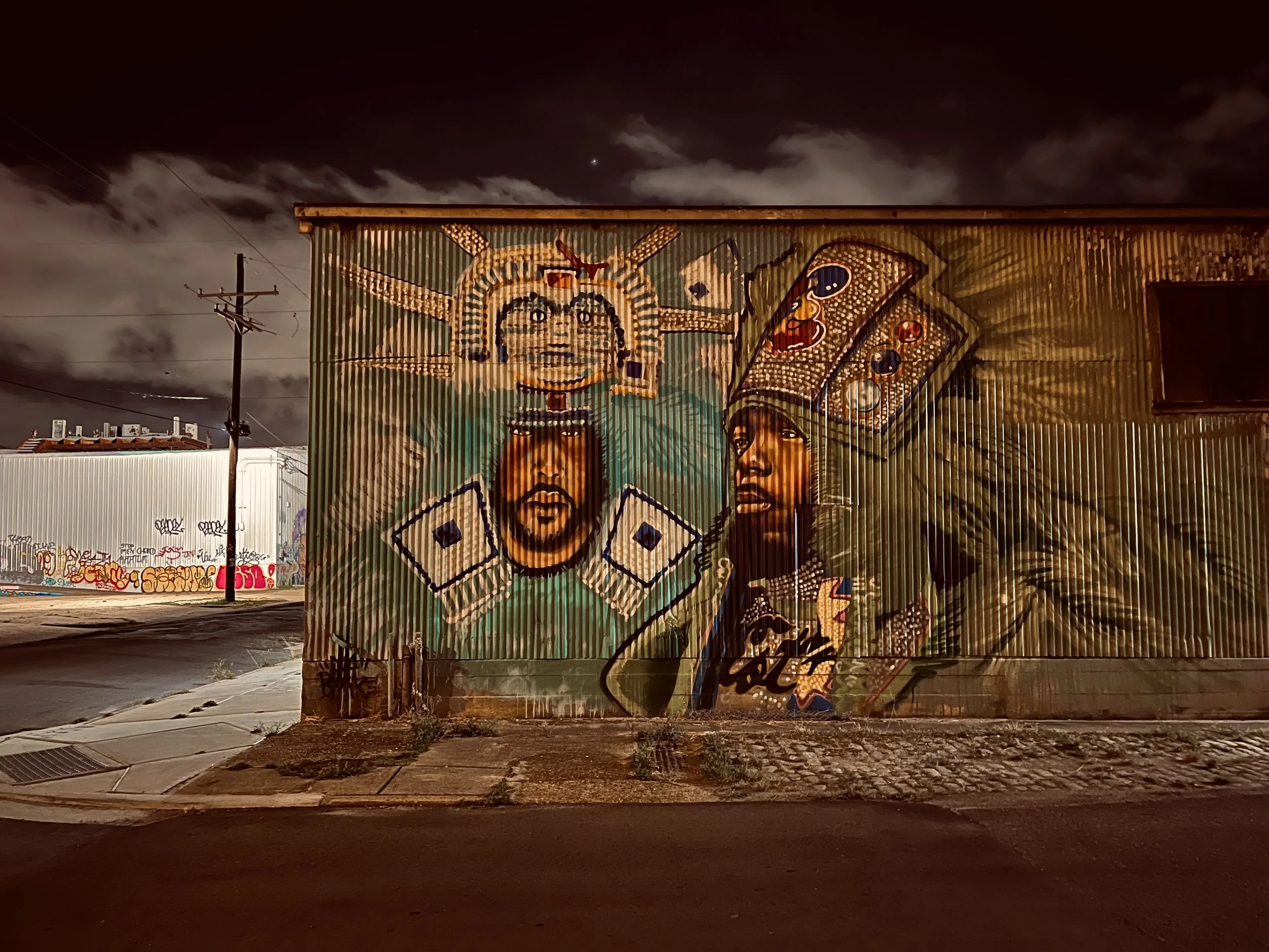 A mural painted on a corrugated metal building at night featuring two stylized faces with headdresses, one with a crown and the other with a headband, using vivid colors and outlines.