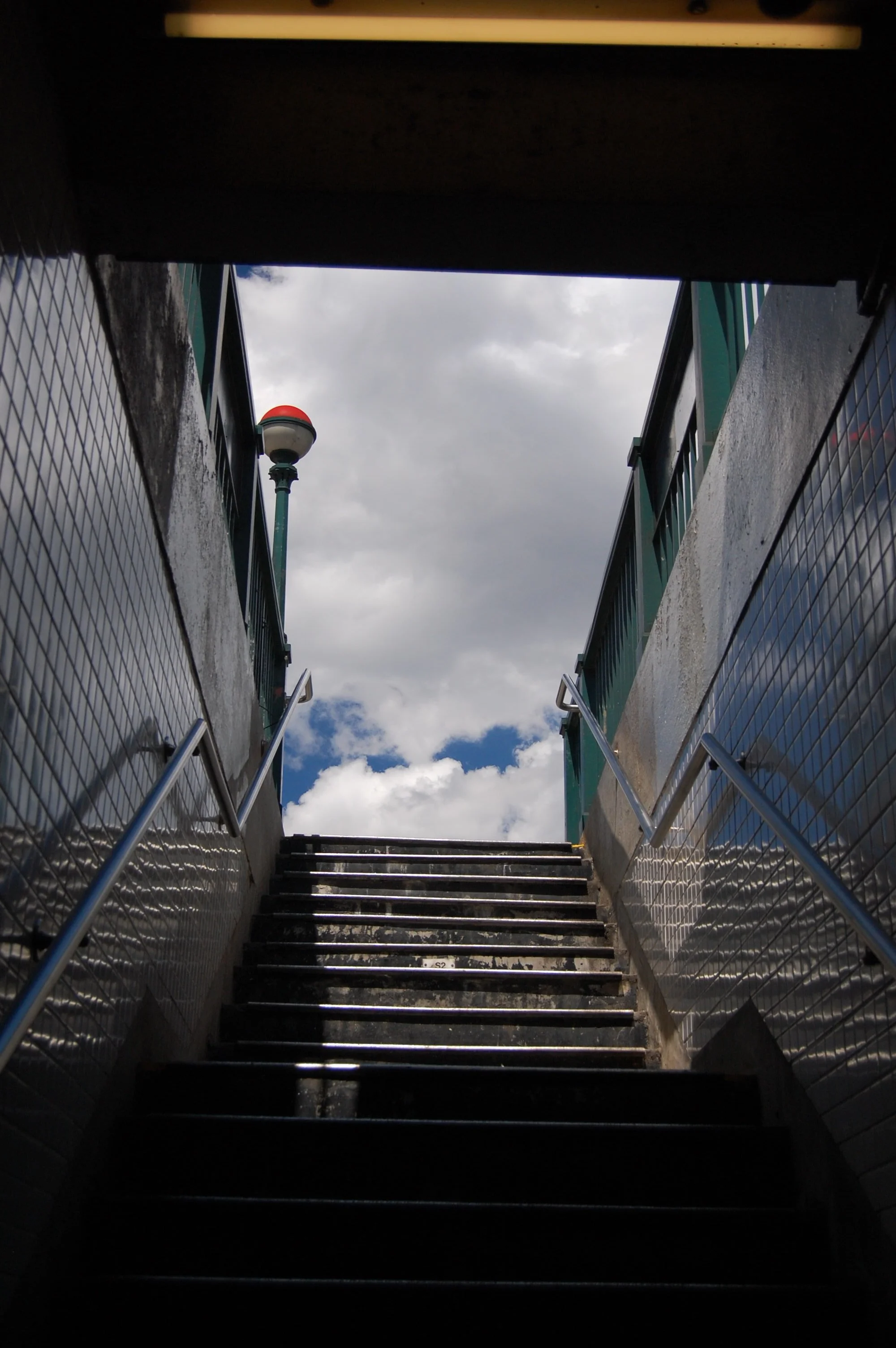 A stairway leading up to an outdoor area with cloudy sky and a lamppost visible in the background, flanked by metal railings on both sides.