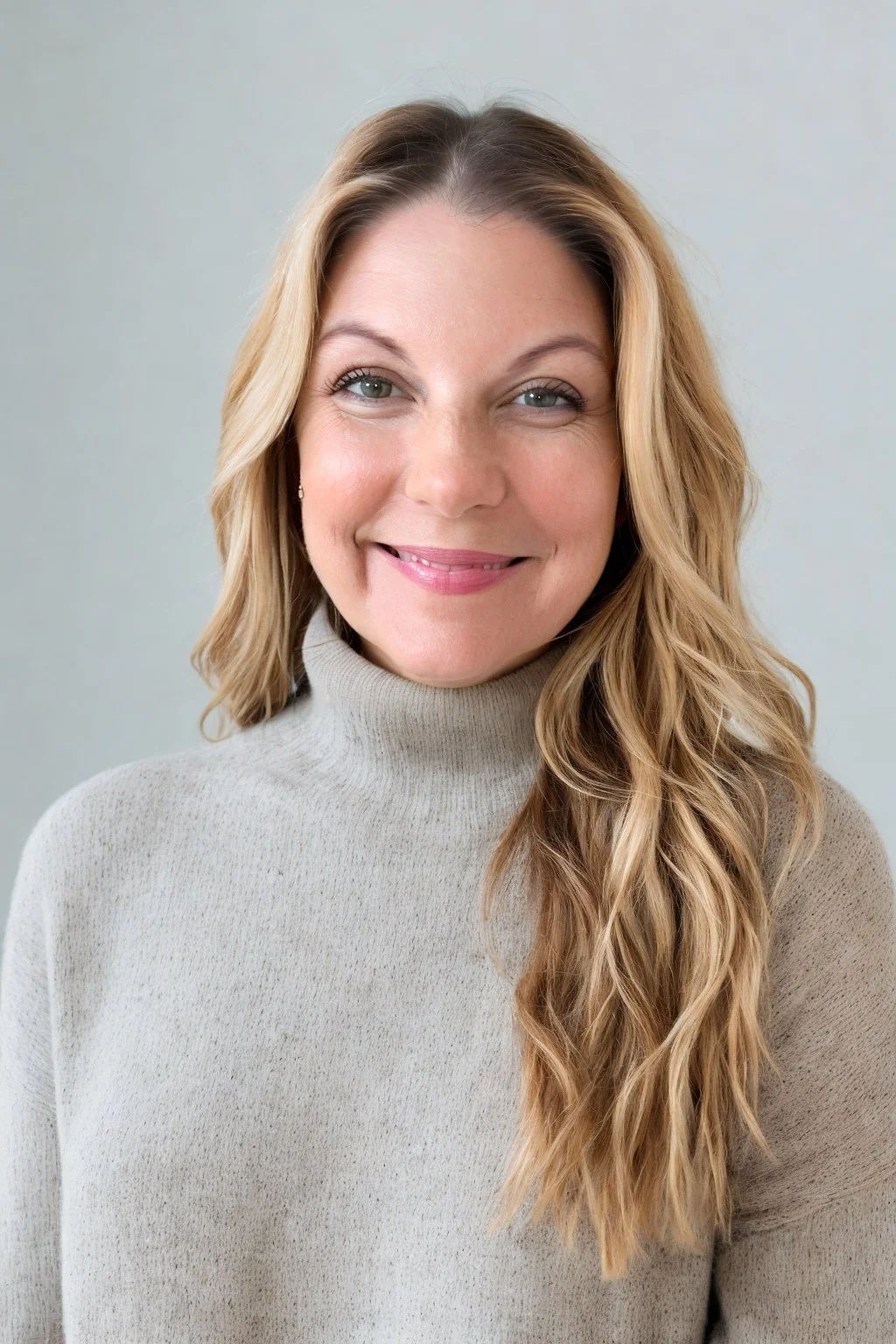 A woman with long blonde hair wearing a light beige turtleneck sweater, smiling, in front of a plain light gray background.