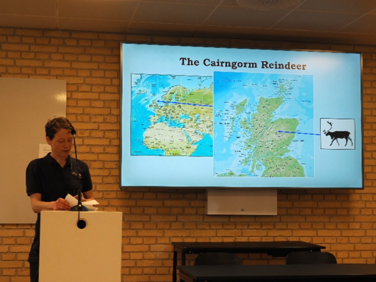 Reindeer herder Andi stands at a podium in front of a powerpoint presentation. The screen shows a map of where the Cairngorm reindeer herder are located.