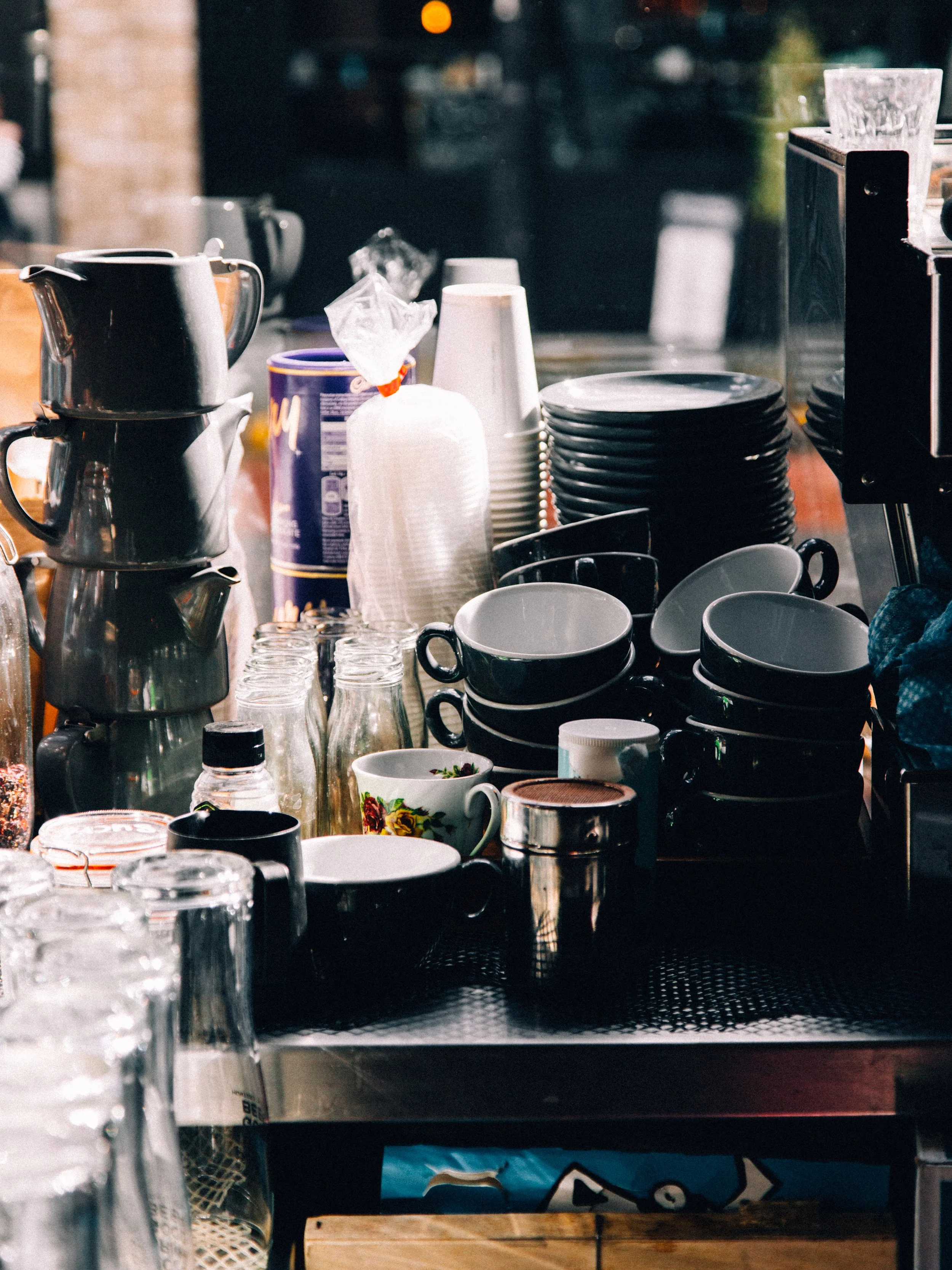 A cluttered bar or cafe counter with stacked black and white coffee mugs, small glasses, a floral mug, foil-covered containers, a pile of paper napkins, and various kitchen items, with bright sunlight reflecting off the surfaces.