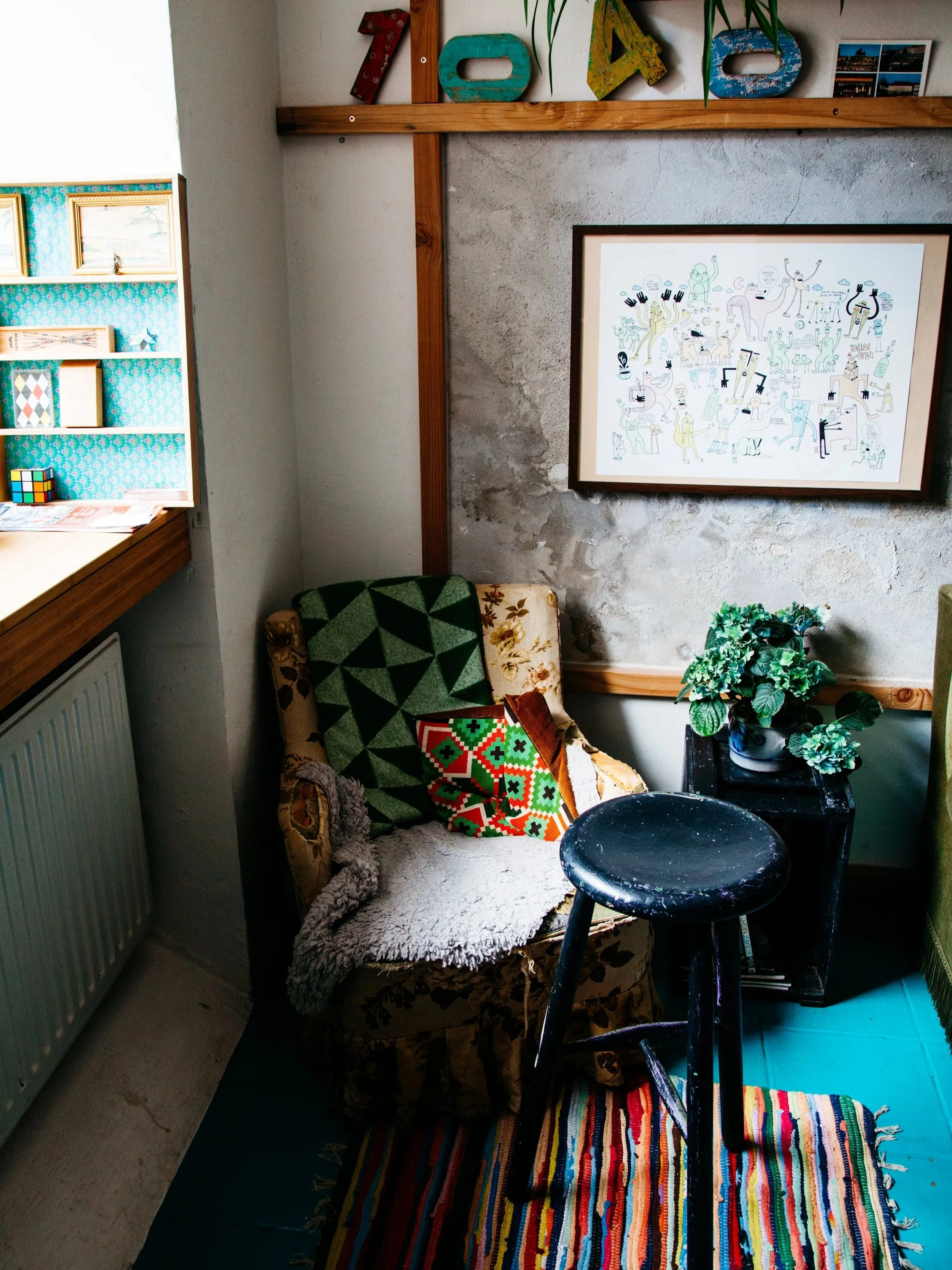 A cozy corner with a vintage armchair covered with a cozy blanket, a colorful embroidered cushion, and a green patterned quilt draped over it. A black stool is in front of a small black table holding a potted plant. A large framed artwork features wh