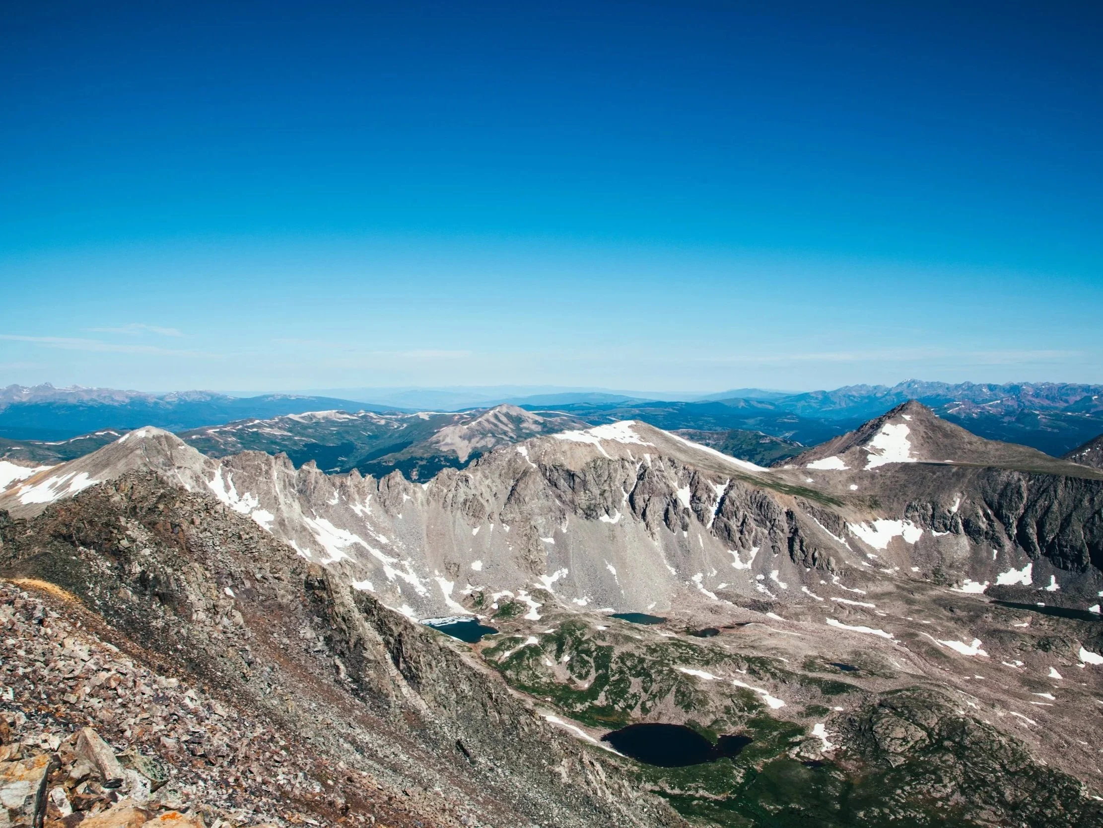 Mountain landscape with rugged peaks, patches of snow, and small lakes, under a clear blue sky.