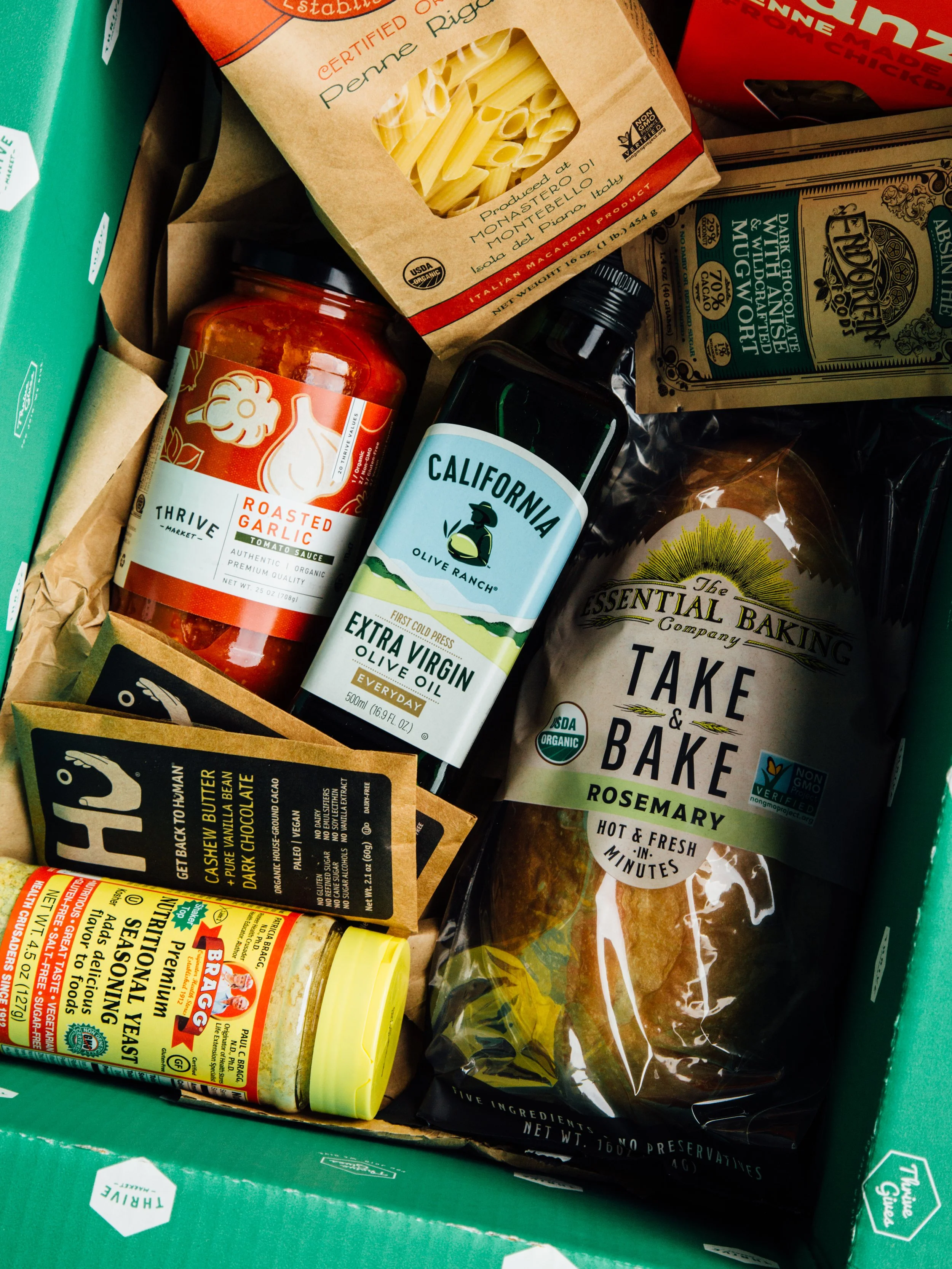 A grocery box contains pasta, tomato sauce, olive oil, a loaf of rosemary bread, a jar of roasted garlic, a box of dark chocolate, and seasoning.