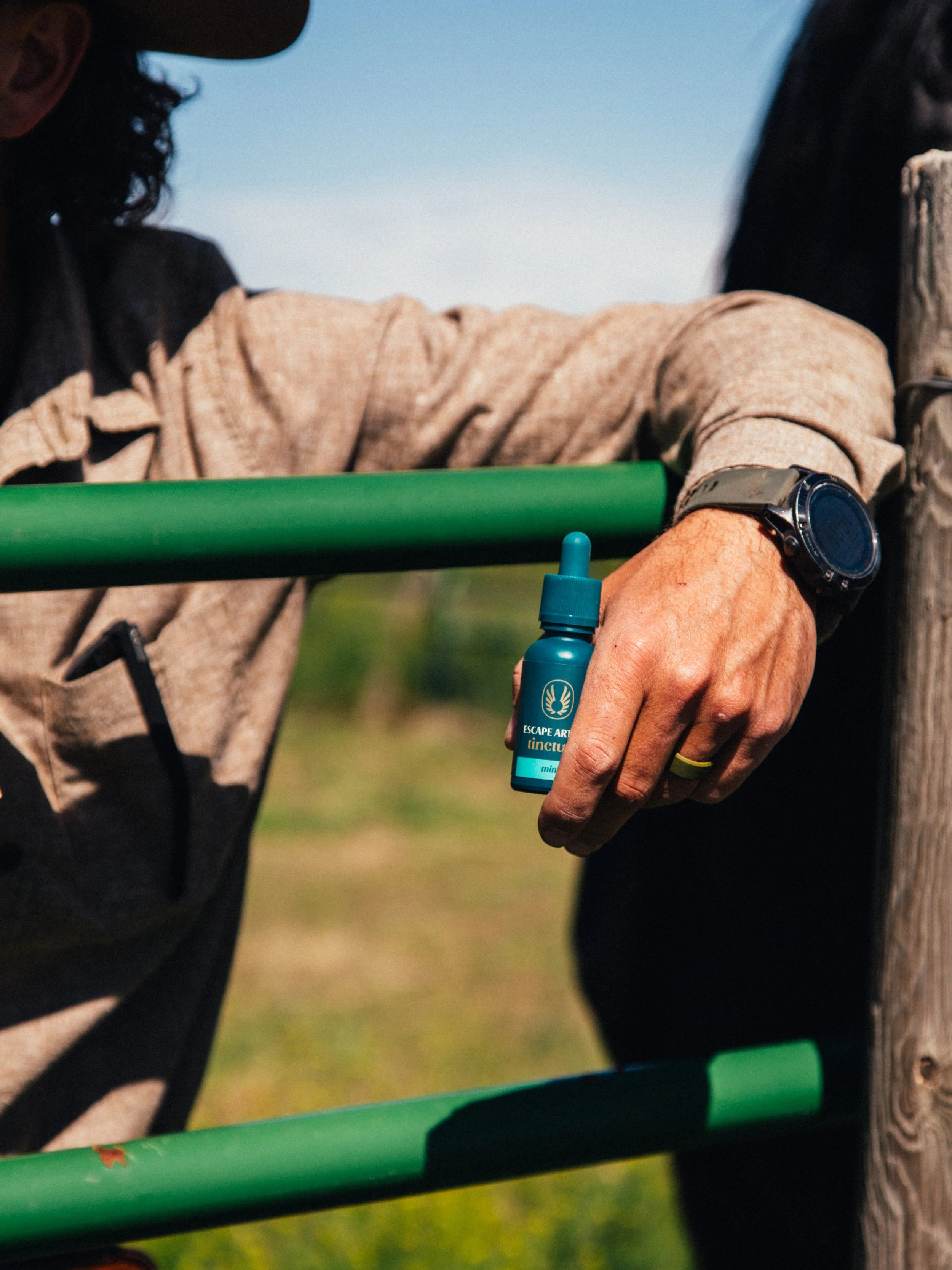 A person holding a small blue bottle of insect repellent outdoors with a green railing in the background.