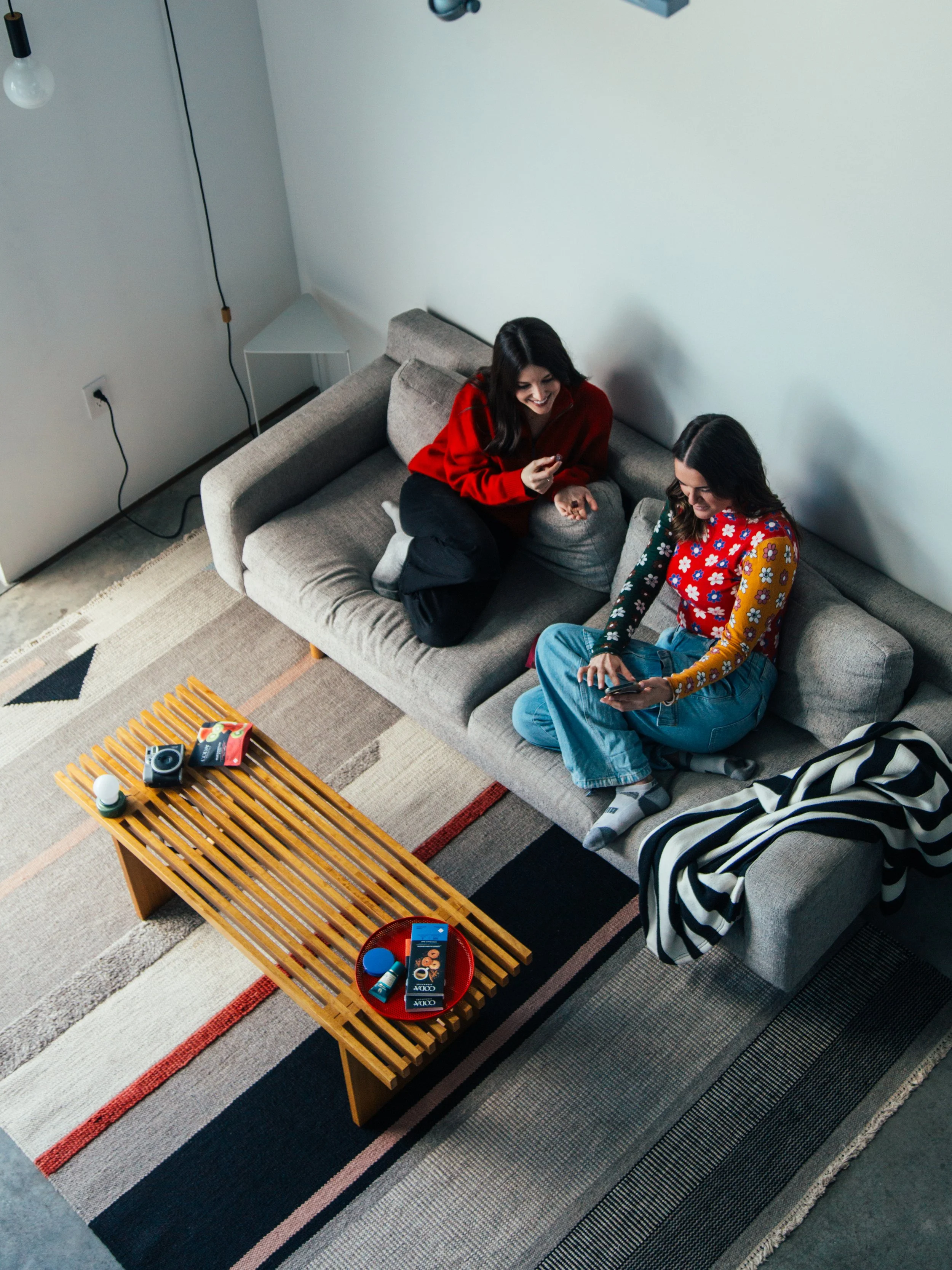 Two women sitting on a gray sofa in a living room, using their phones and smiling, with a wooden coffee table in front of them holding various items, and a rug underneath.