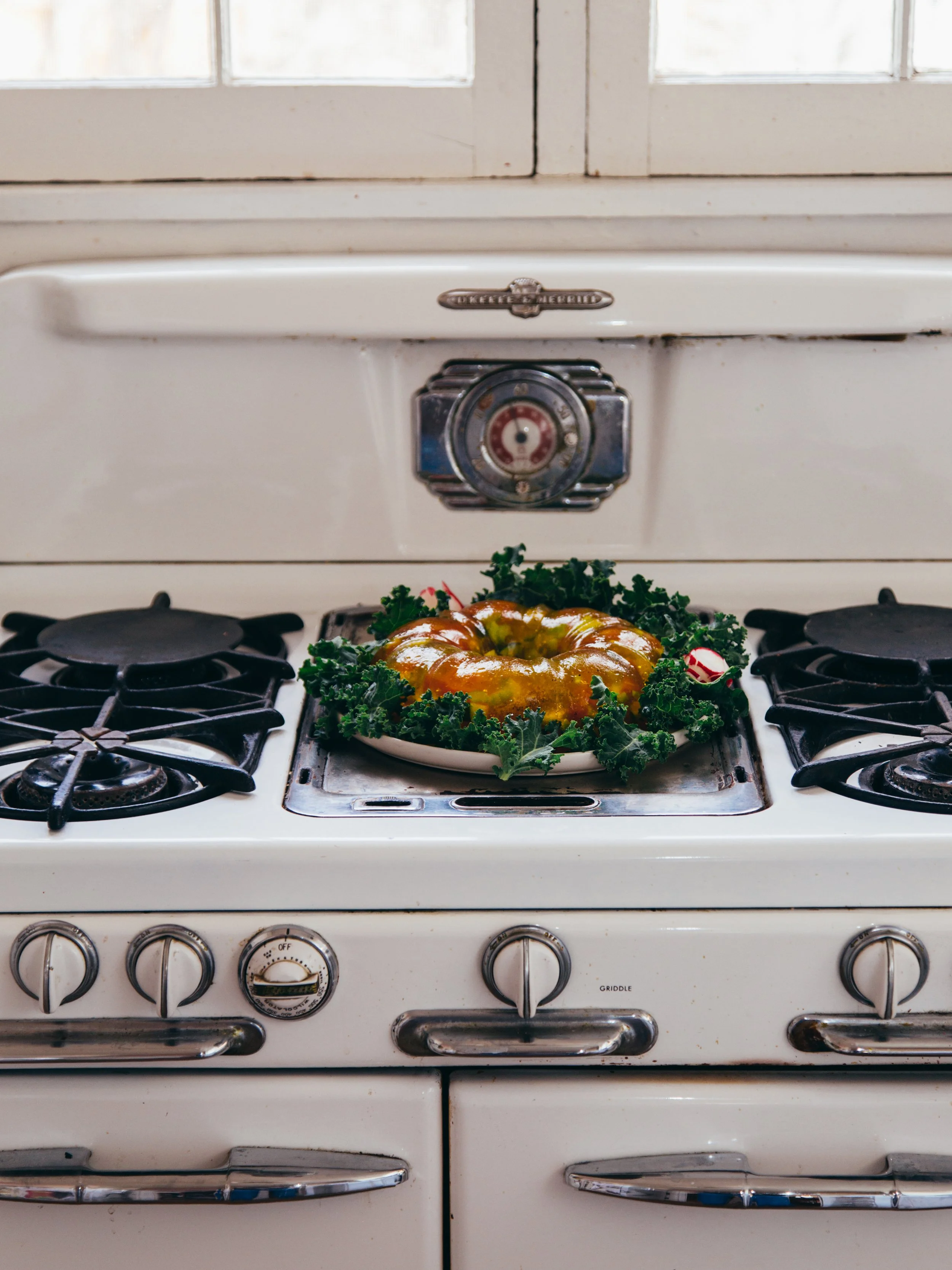 A vintage white gas stove with a decorative metal emblem above the oven door, a circular control dial, and four burners. A green decorated holiday wreath with a yellow glazed bundt cake is placed on the stove.