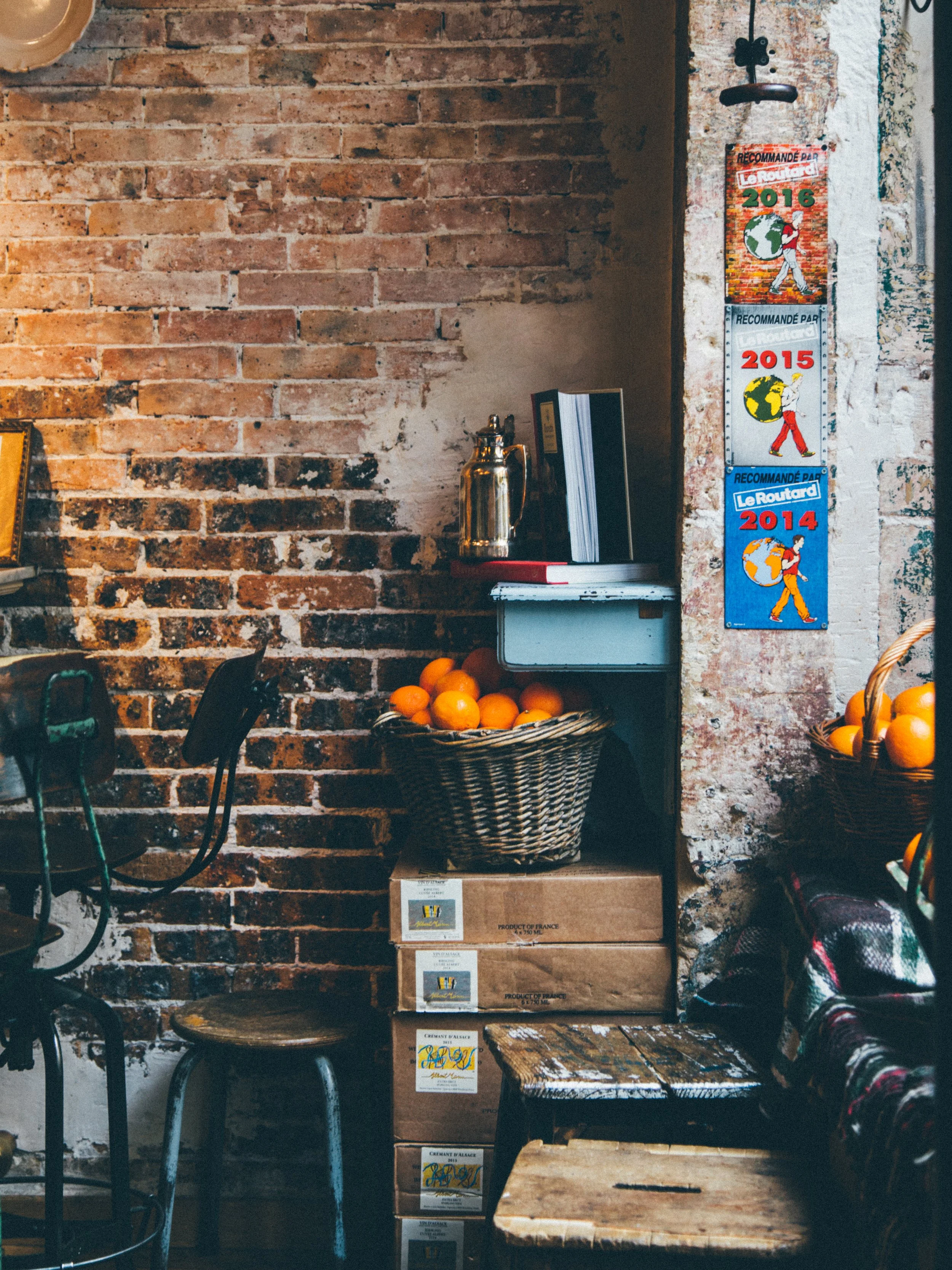 A rustic interior scene with a brick wall and a corner featuring a wicker basket of oranges, stacked boxes, and a small table. There are some books, a metal thermos, and advertising posters on the wall.