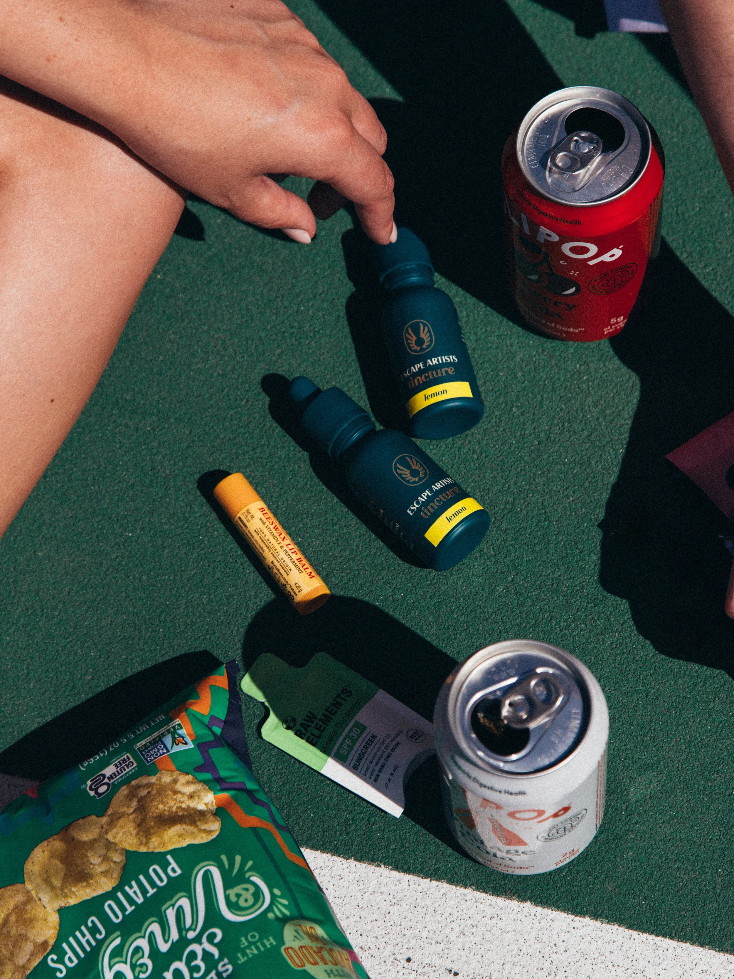 A person sitting on a green surface with two opened cans of soda, a couple of essential oil bottles, a tube of lip balm, a green ticket, and a bag of potato chips next to them.