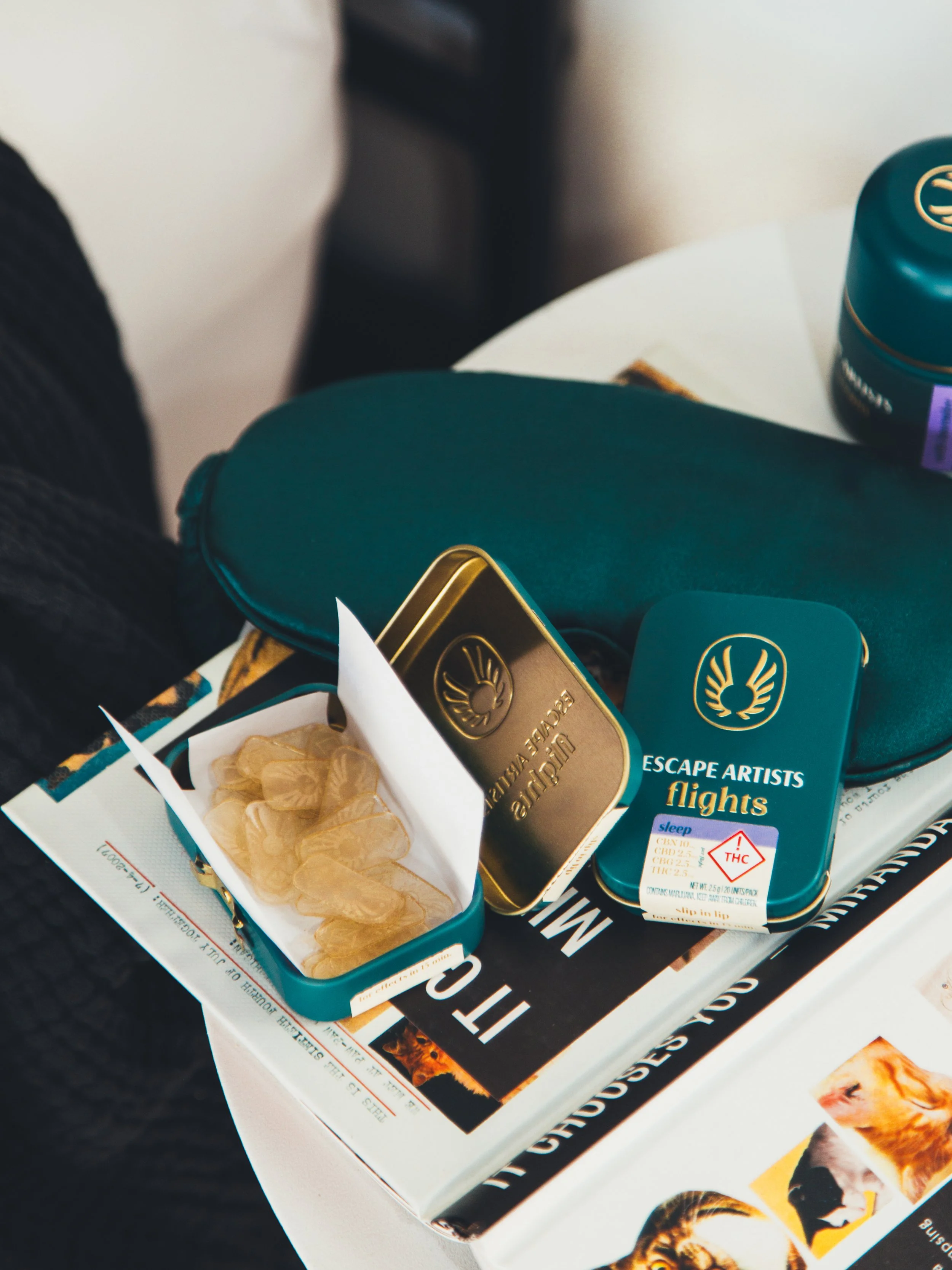 Open teal tin container with cannabis gummies, another teal container labeled "Escape Artists Flights," and a teal case on a table with a newspaper underneath.