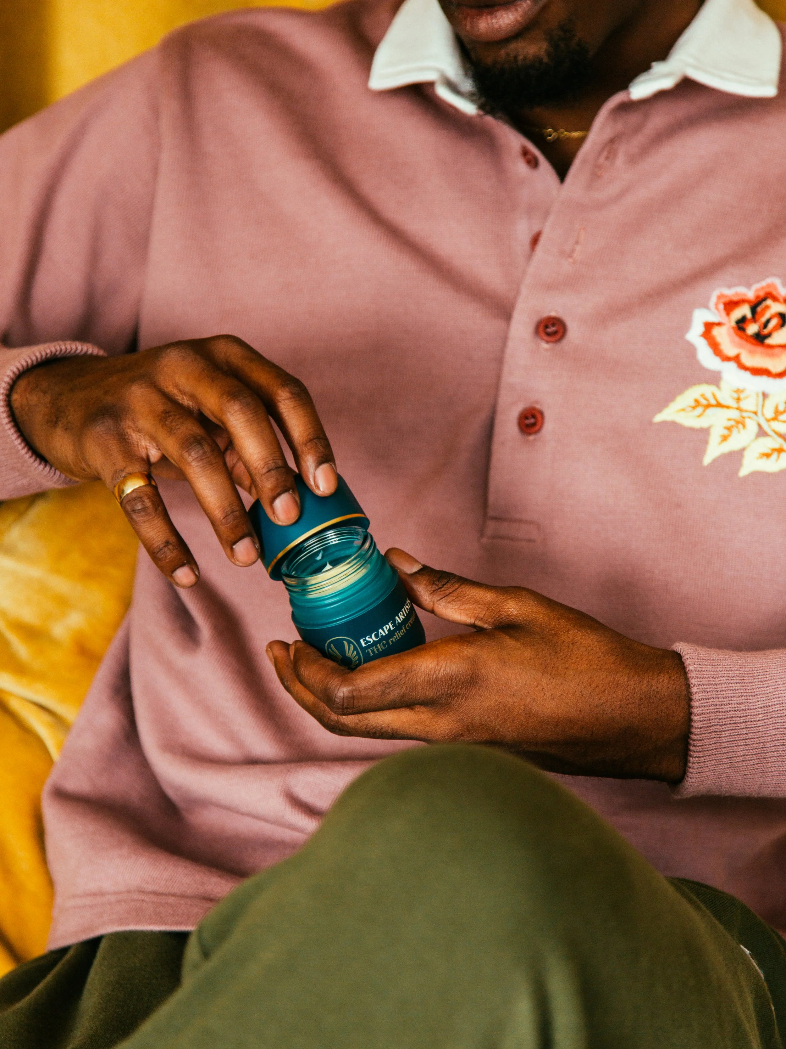 A person is opening a teal container labeled "Escape Arms THC relief" while sitting on a yellow couch, wearing a pink shirt with floral embroidery on the chest.