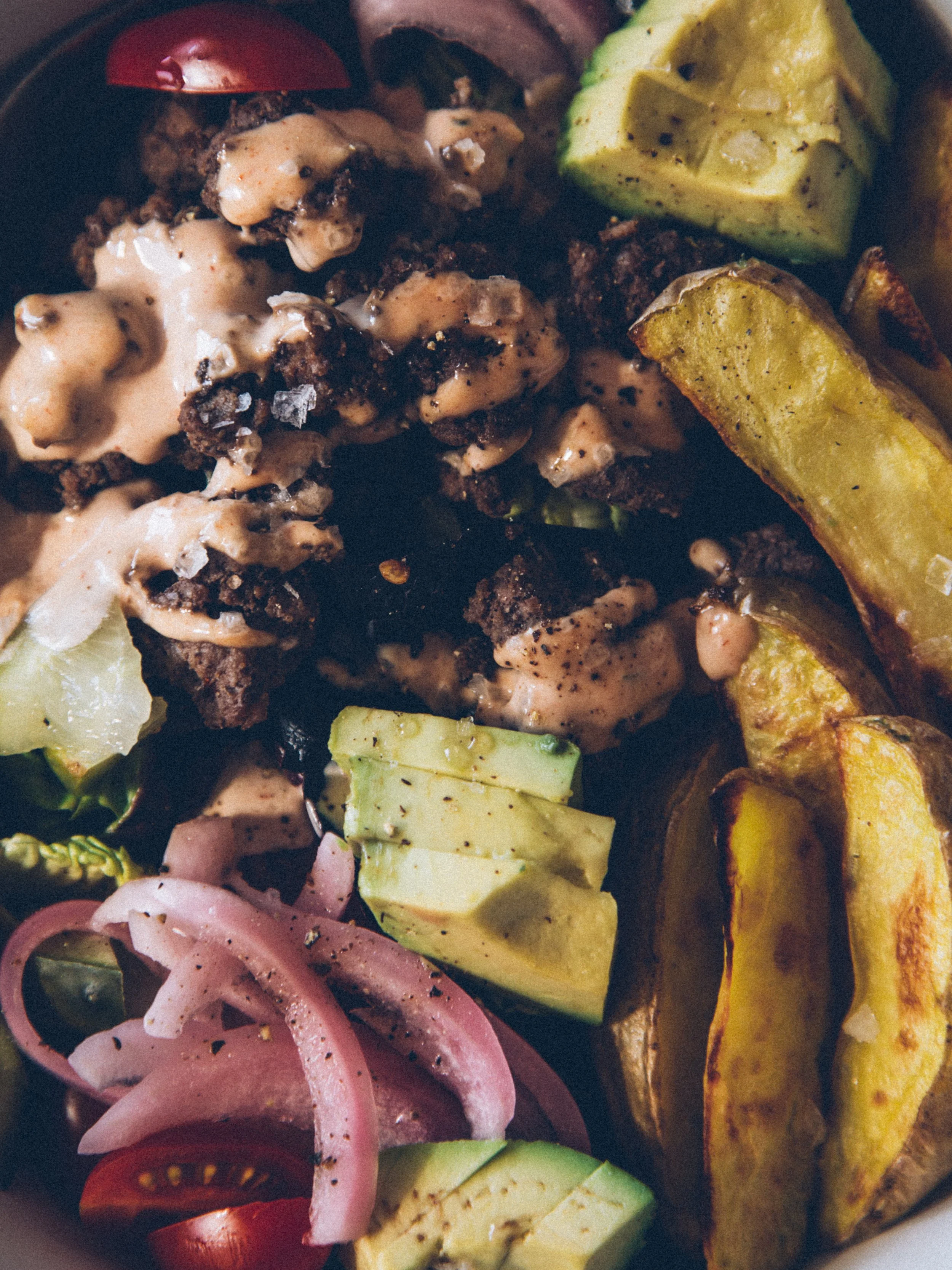 A plate of food with chunks of grilled meat topped with sauce, slices of avocado, roasted sweet potatoes, and a salad with pickled onions and cherry tomatoes.