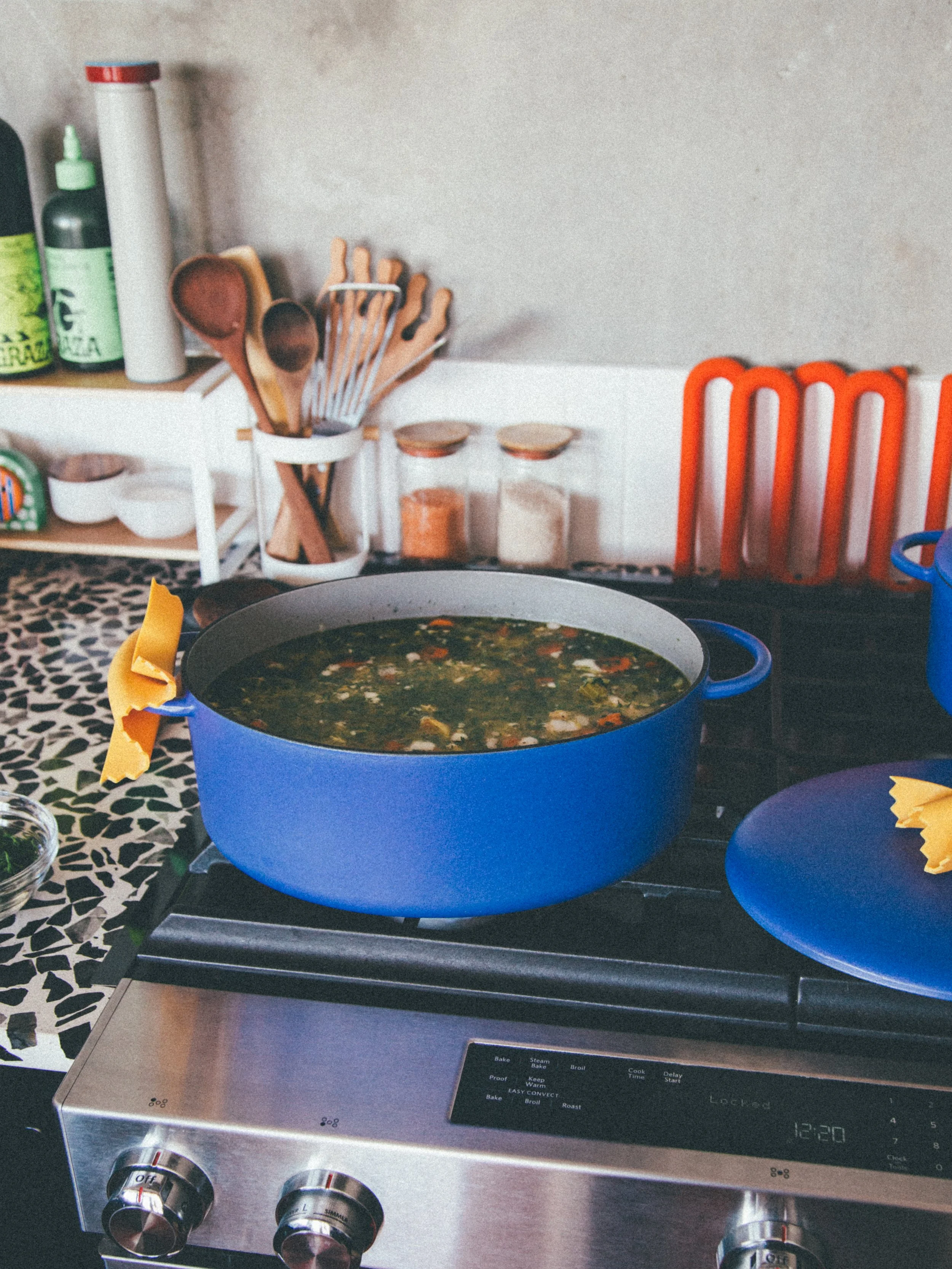 A blue pot on a stove filled with soup or stew with vegetables and meat pieces. There are yellow napkins folded on blue plates around the stove. In the background, a kitchen counter holds utensils, spices in jars, and bottles.