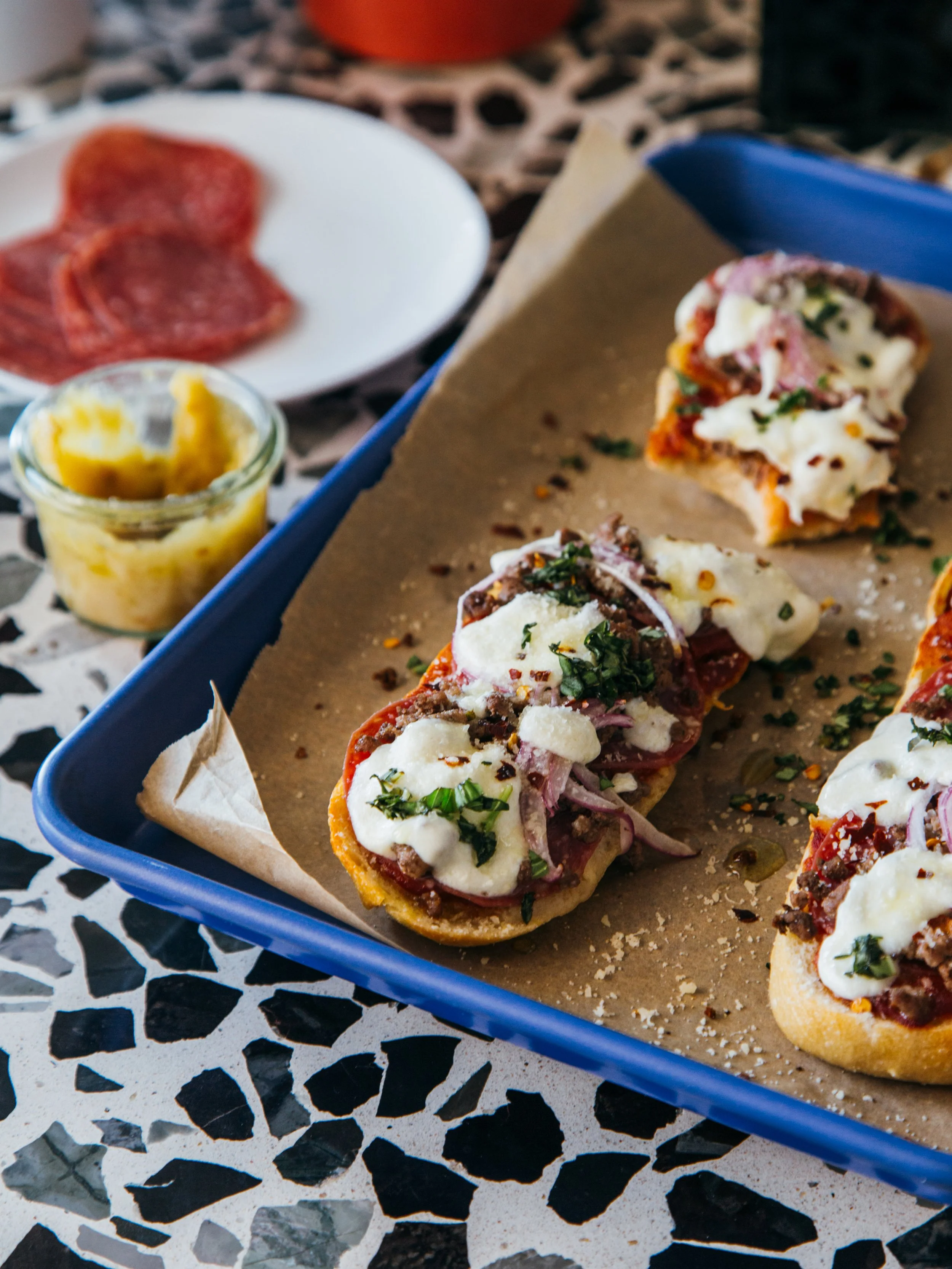 Slices of mini pizza topped with cheese, herbs, and possibly meats on a baking sheet lined with parchment paper.