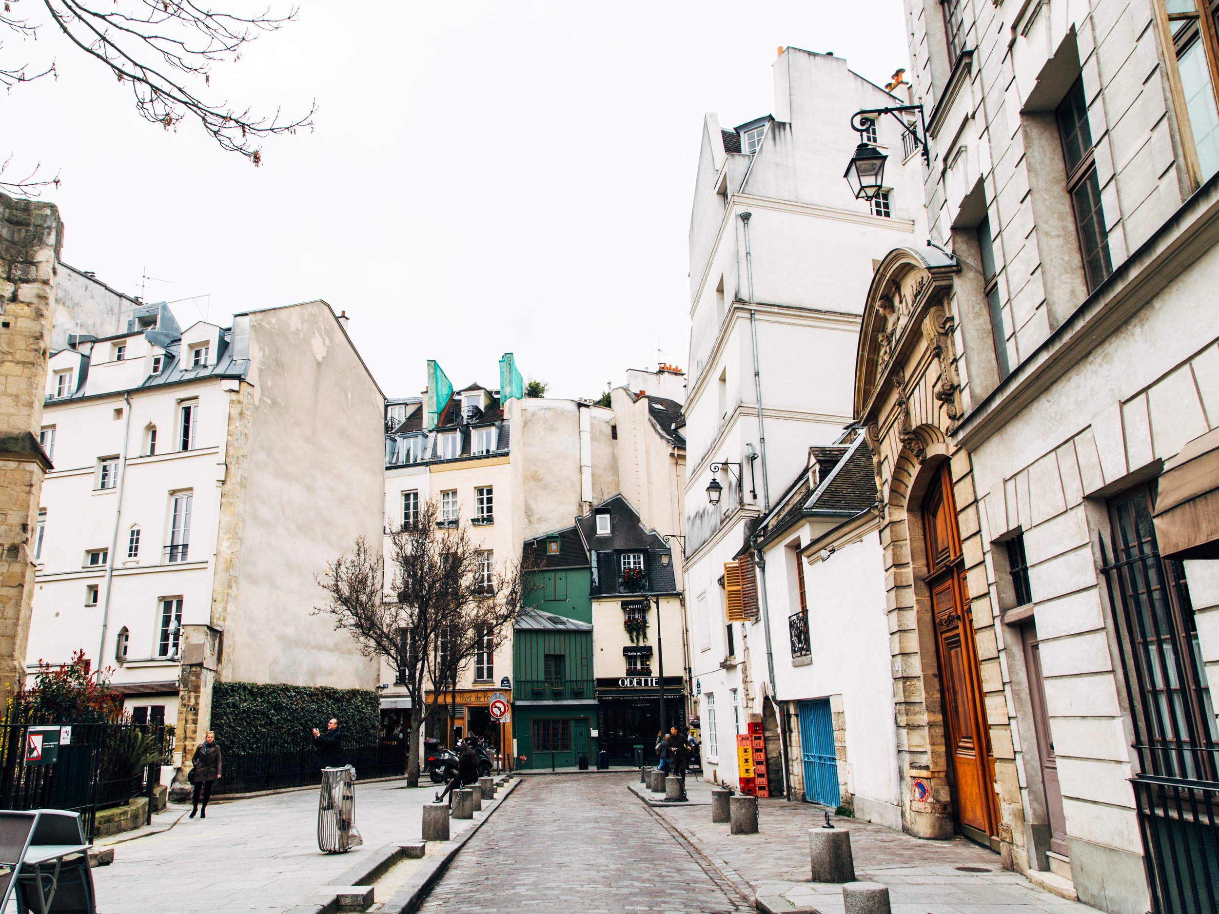 A narrow cobblestone street in a European city with old buildings on both sides, some with shutters and decorative facades, and a few pedestrians walking.