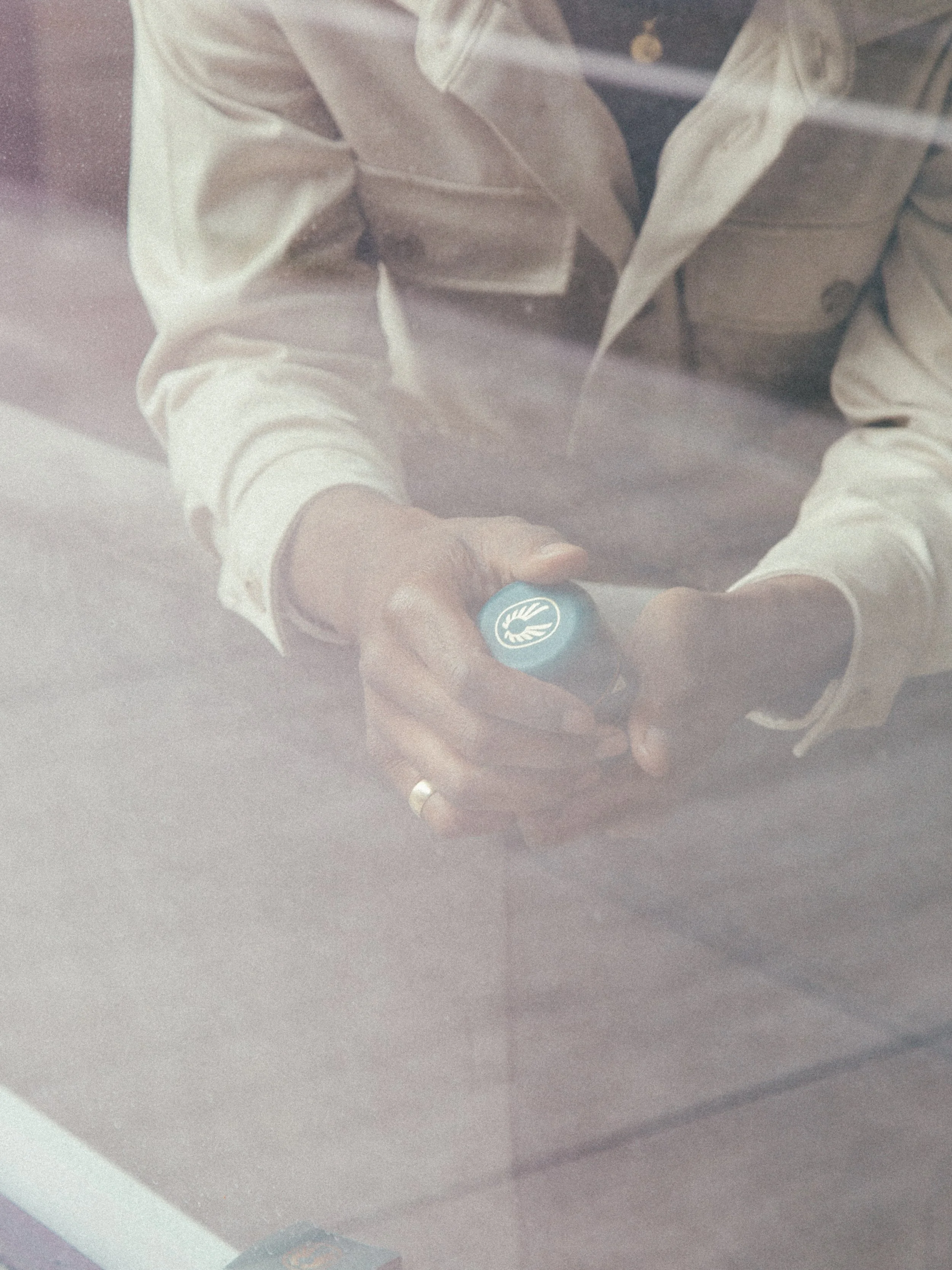 Person wearing a beige jacket holding a blue object with a white leaf logo on top, sitting at a table with a blurred background and a glass reflection in the foreground.