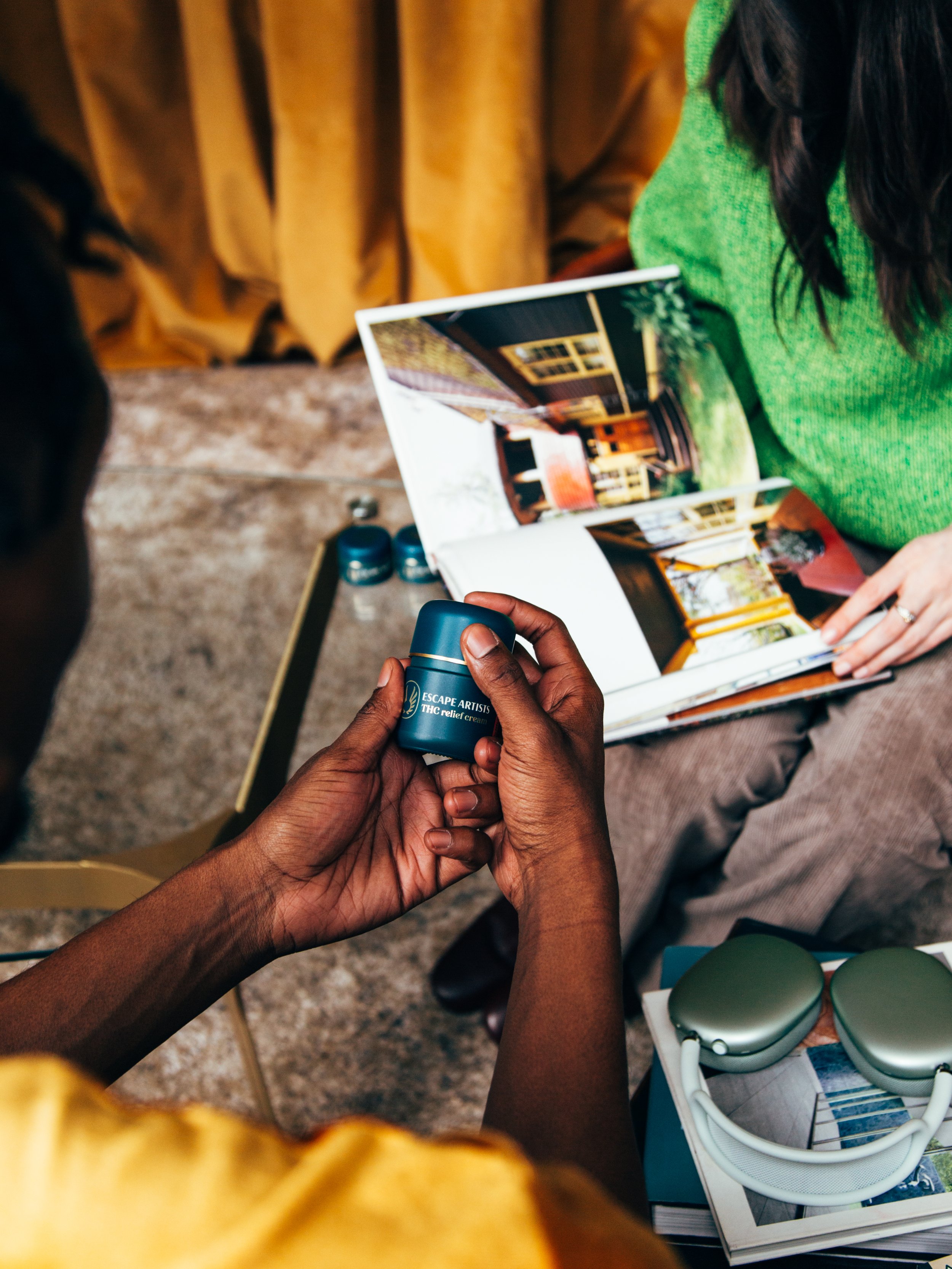 A person holding a small jar labeled 'Escape Artists THC Relief Cream' while another person shows them a photo album with pictures of interior room designs.