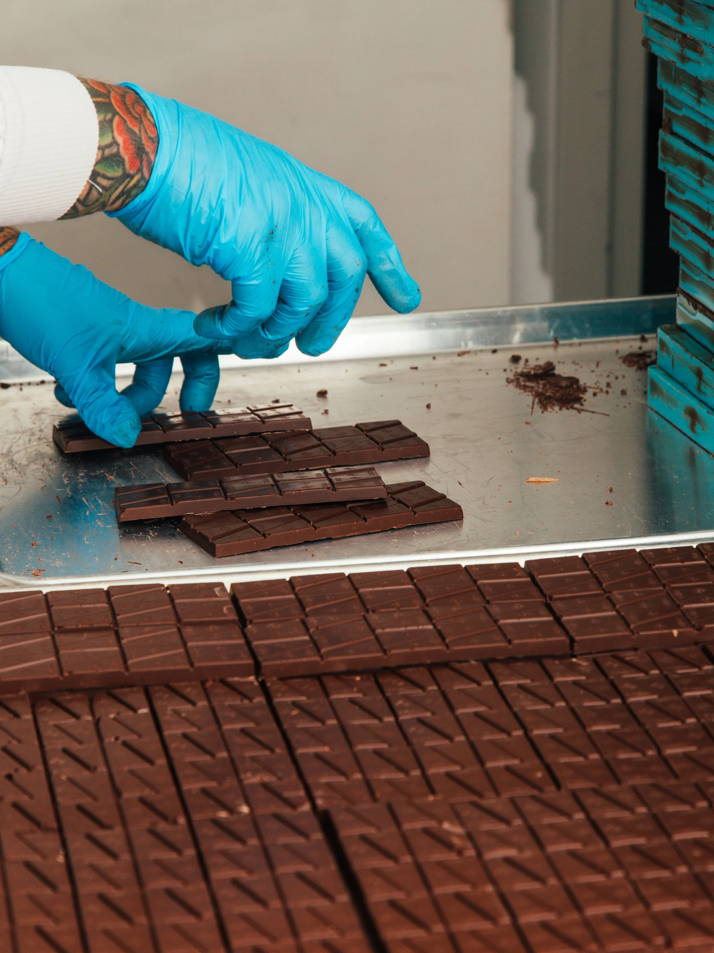 A person wearing blue gloves and white sleeves is handling sections of chocolate on a metal surface, with stacks of chocolate nearby.