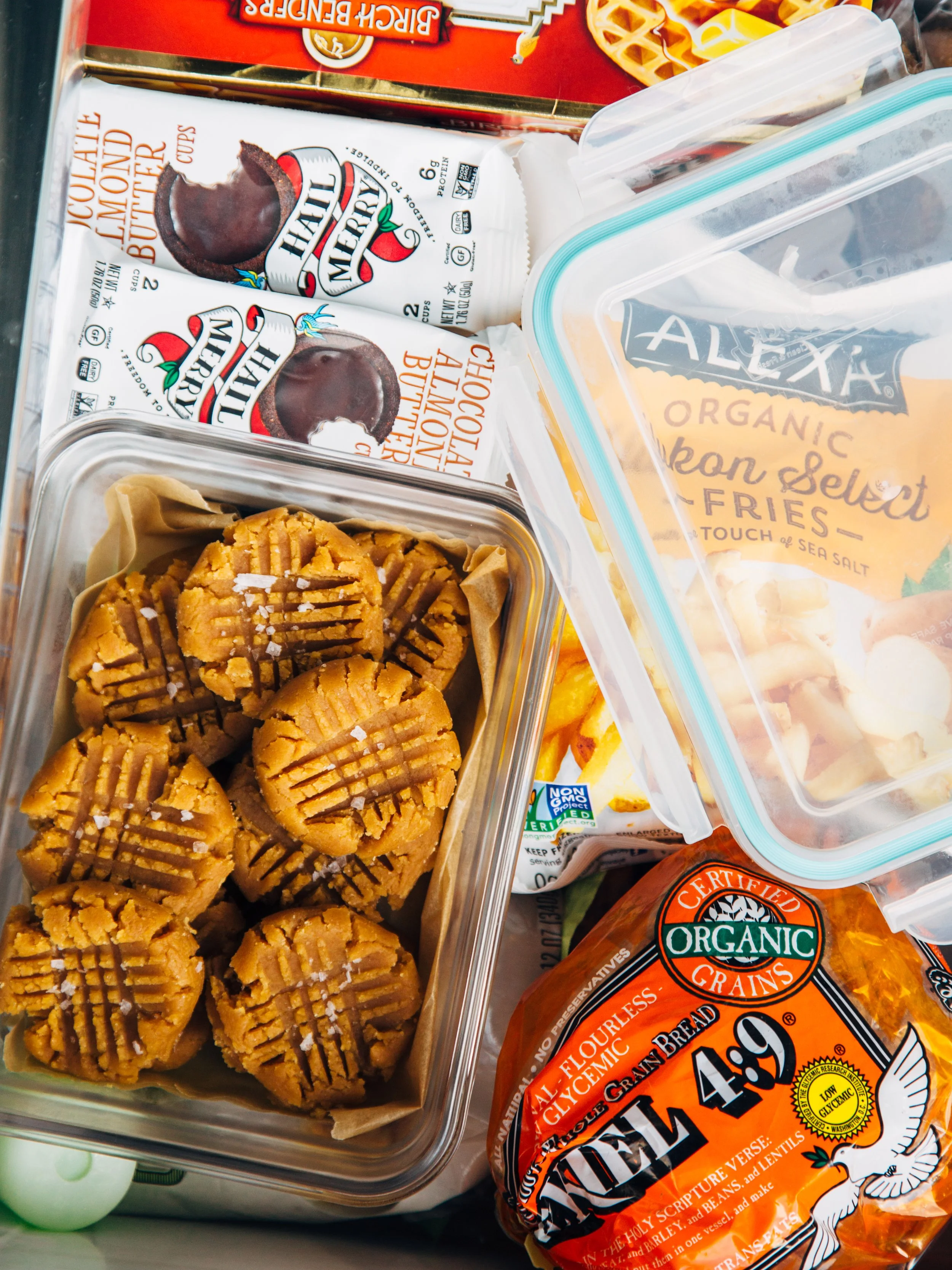 A collection of food items including a container of peanut butter cookies, a box of organic lemon select fries, a loaf of organic gluten-free bread, and some chocolate almond butter packets.