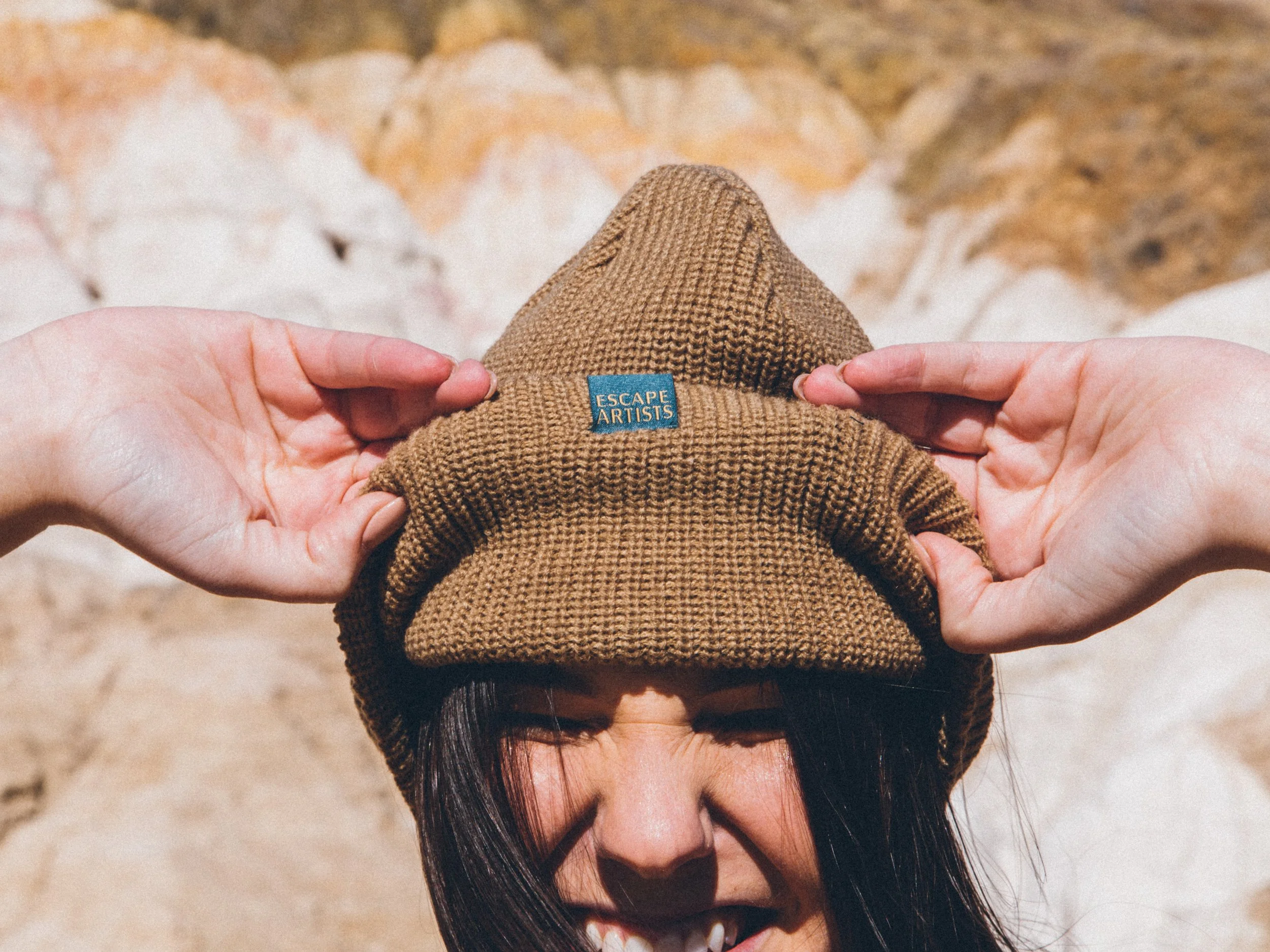 Person wearing a brown knit beanie hat outdoors, holding the hat with both hands, with rocky terrain in the background.