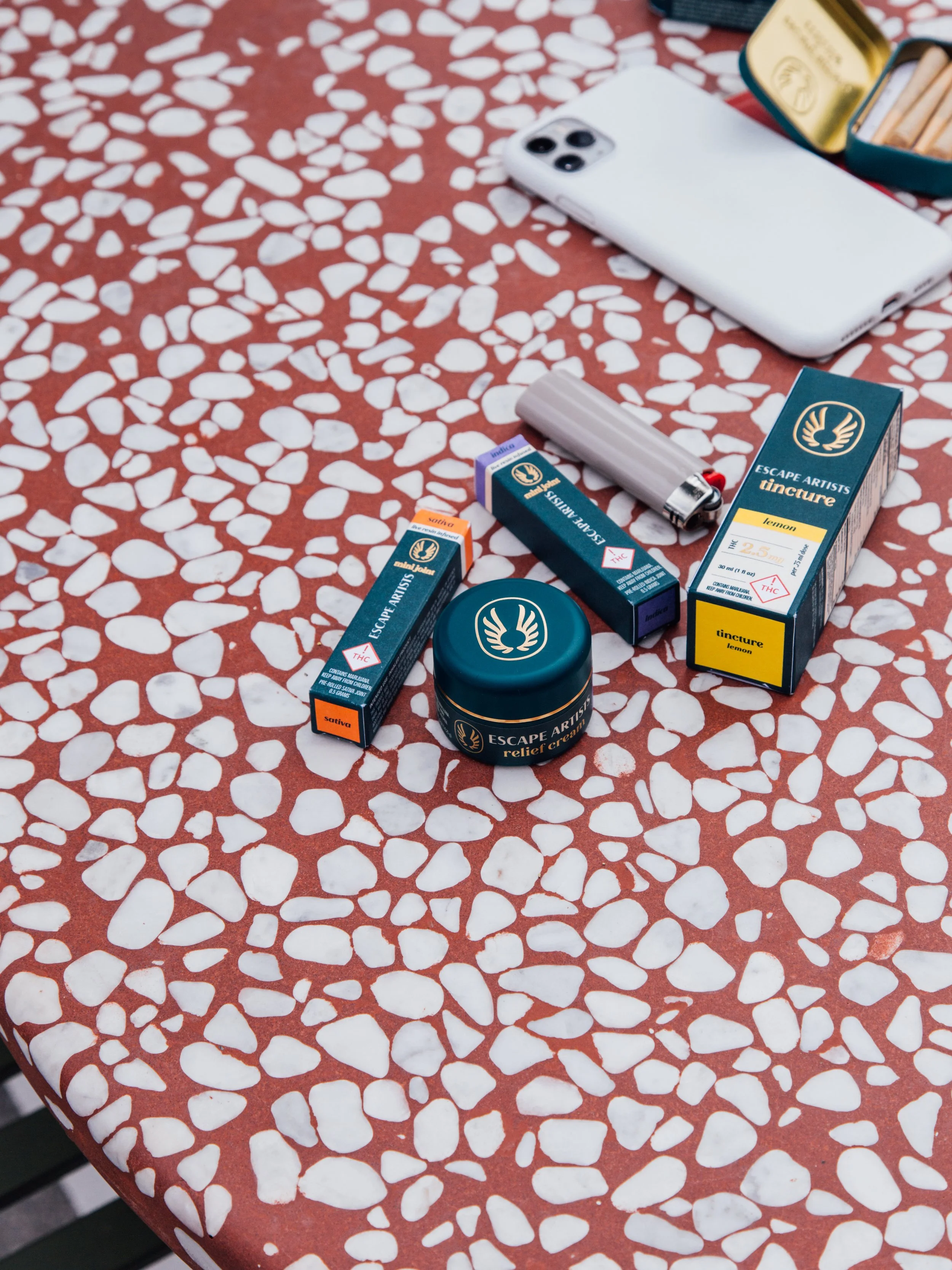 Assorted cannabis-related products and accessories on a red and white patterned table. Items include cannabis relief cream, tincture boxes labeled lemon, lighters, a tin box, and a smartphone.