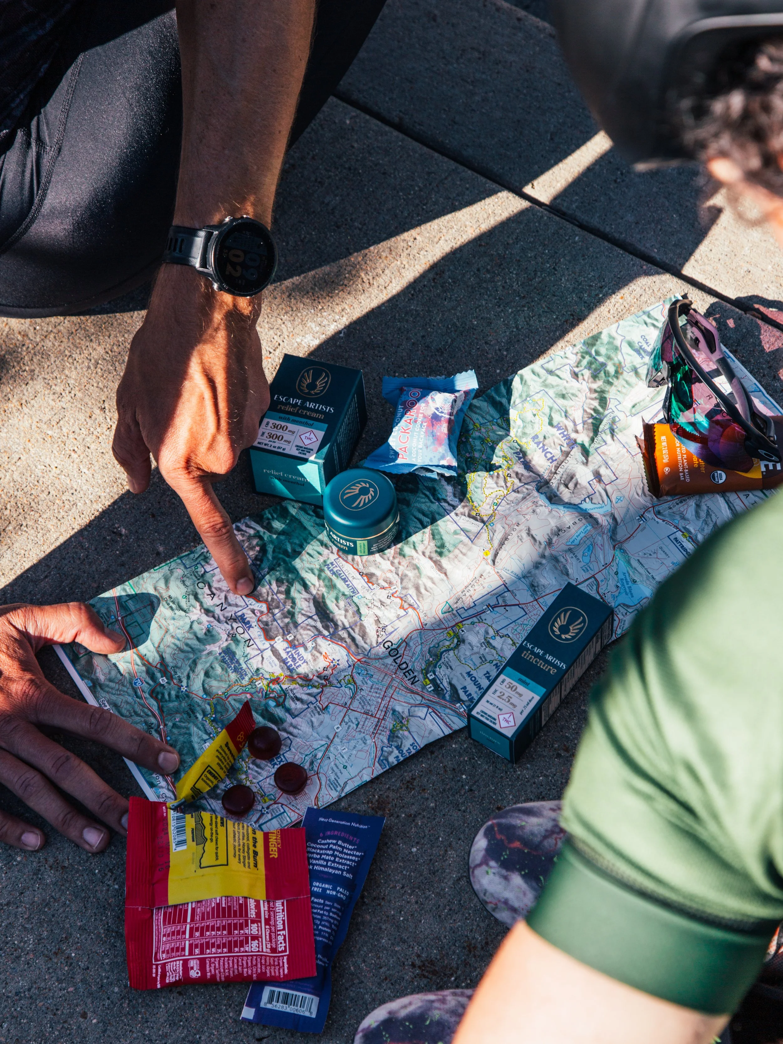 Two people sitting on pavement with a large topographic map spread out between them, surrounded by snacks, sun protection, and medications, possibly planning a hiking or outdoor adventure.