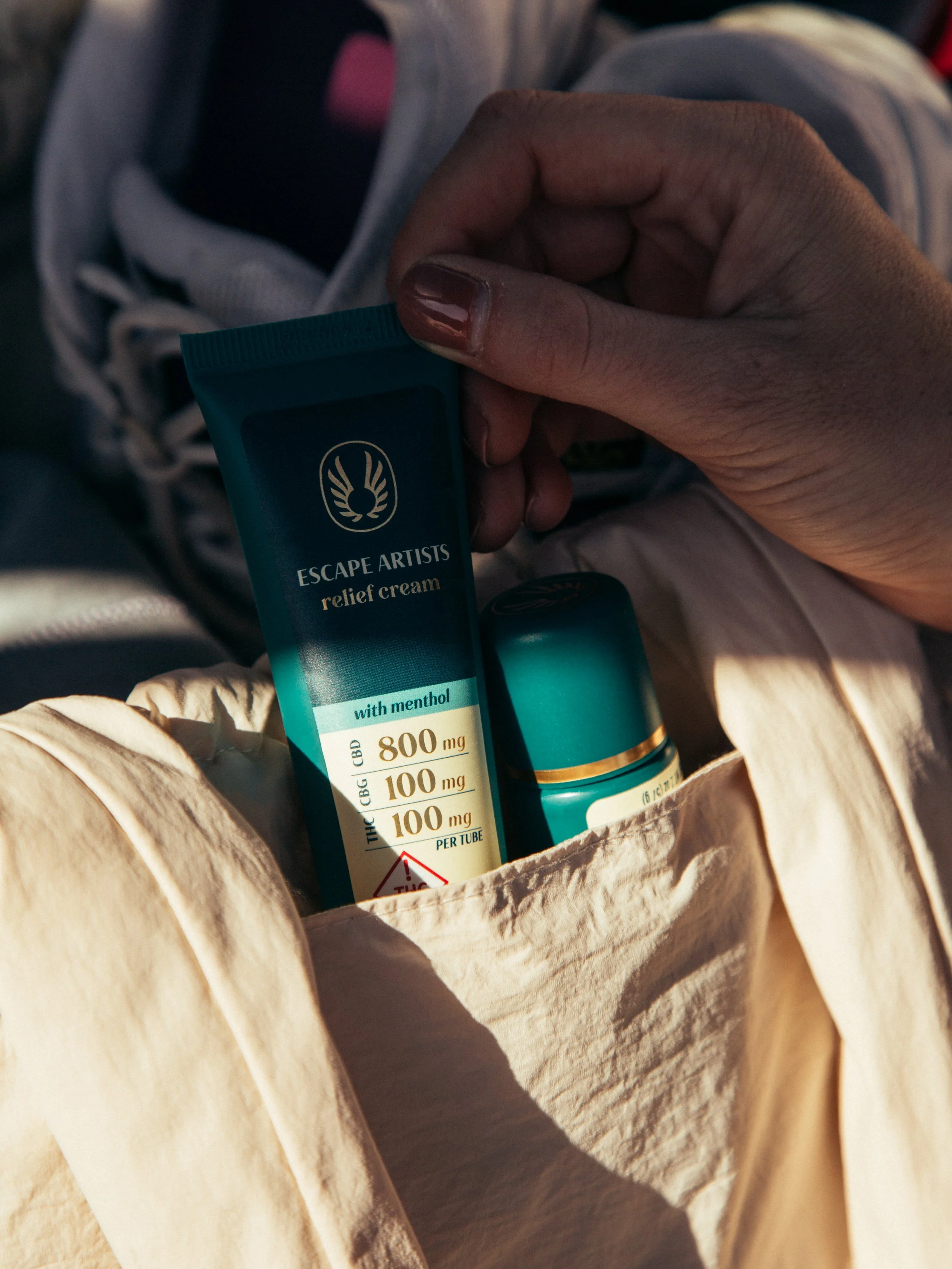 A hand holding a tube of Escape Artists relief cream with menthol, placed in a paper bag along with other items, possibly in a vehicle.