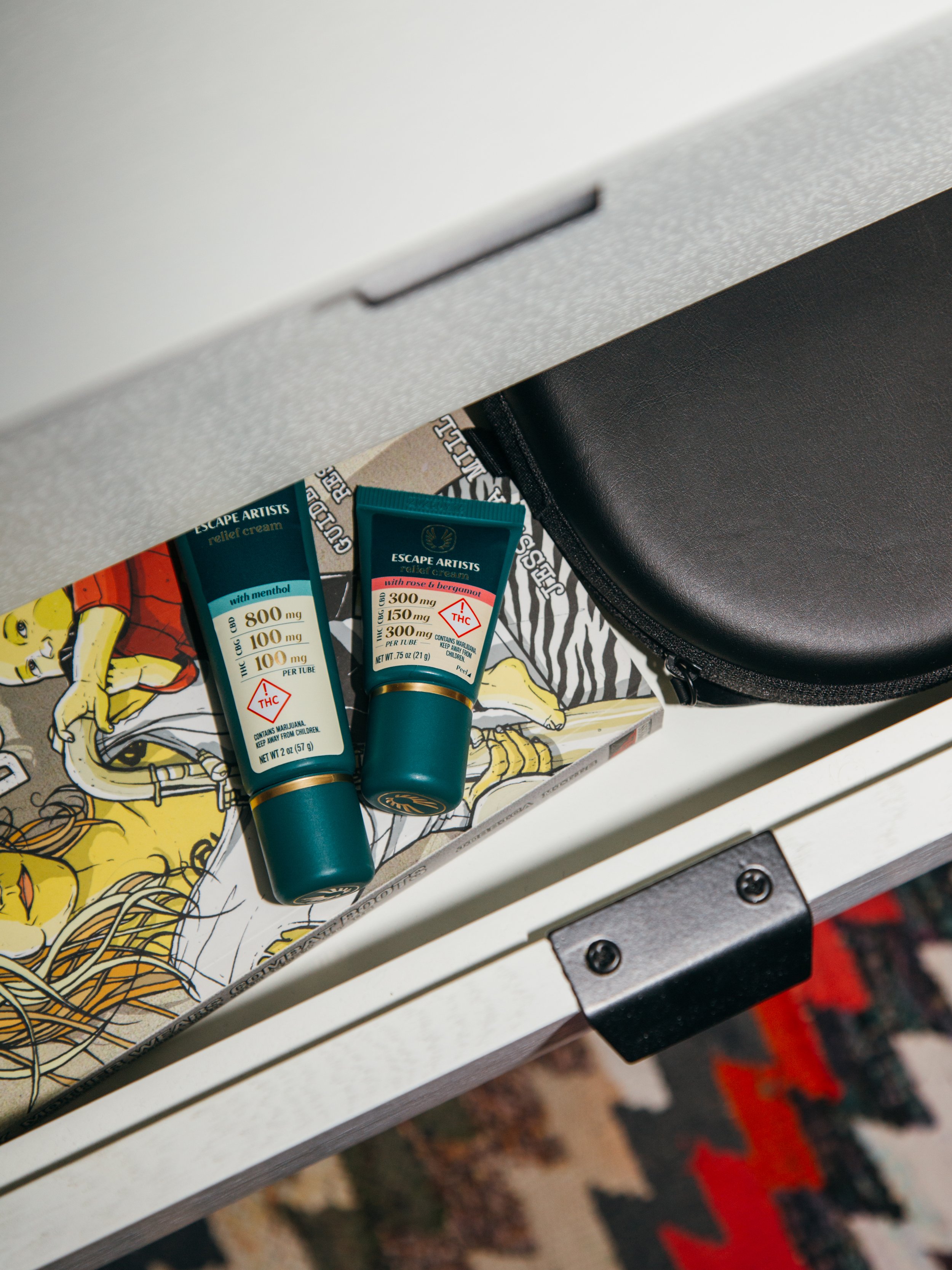 Two tubes of Escape Artists relief cream with marijuana warning labels on a colorful illustrated surface, partially hidden under a desk drawer.