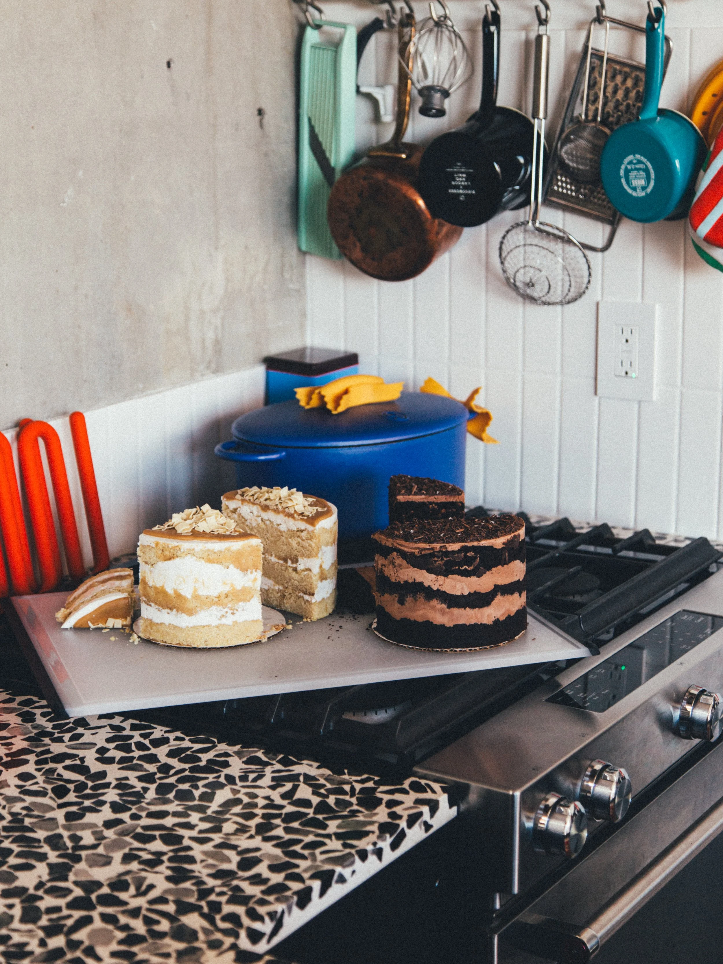Two decorated cakes on a white cutting board on a kitchen stove, with kitchen utensils and containers hanging on the wall in the background.