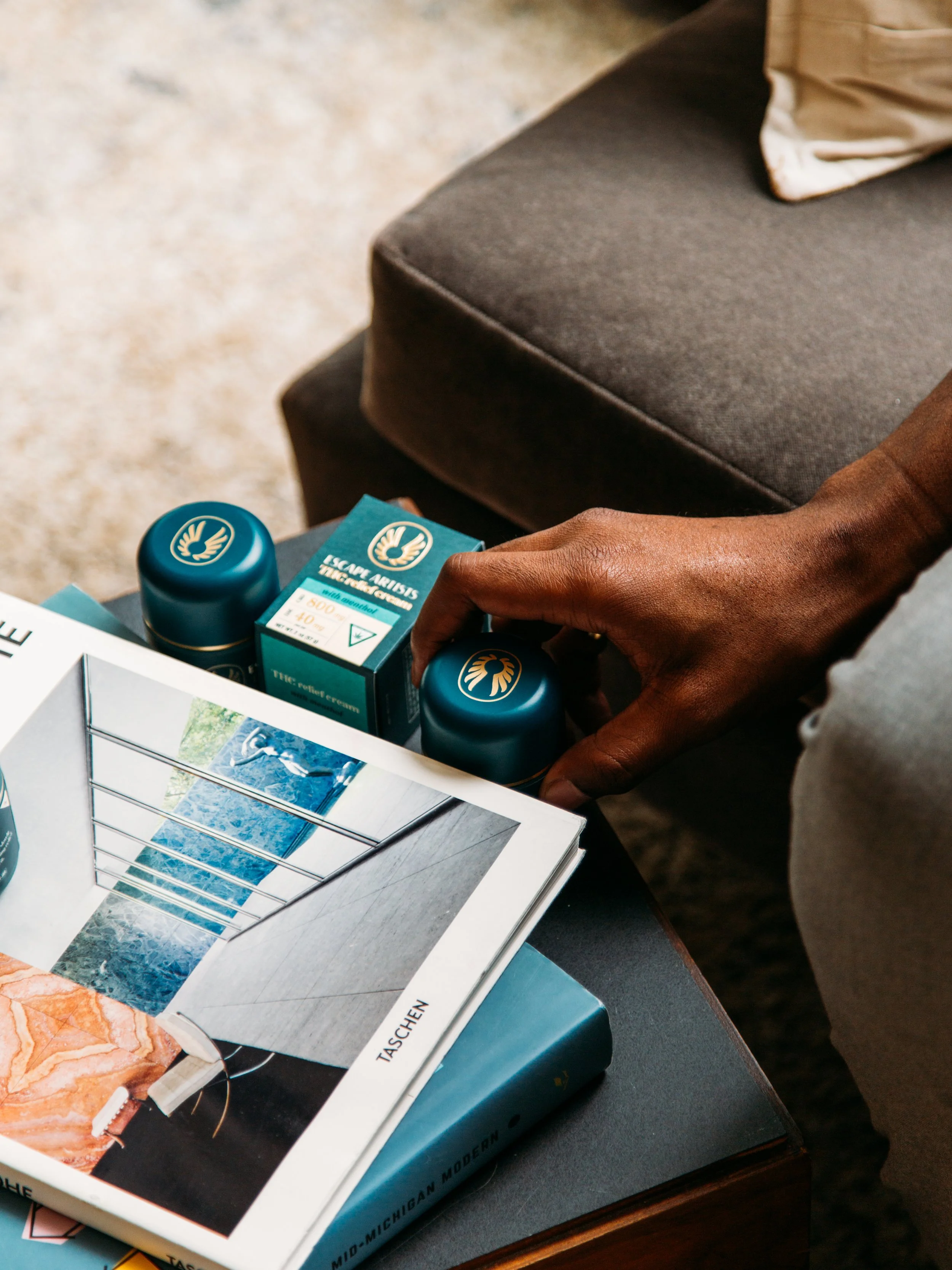 A person with dark skin reaching to pick up two teal jars with golden logo, on a dark brown table near a gray sofa, with travel books and a magazine accessible.