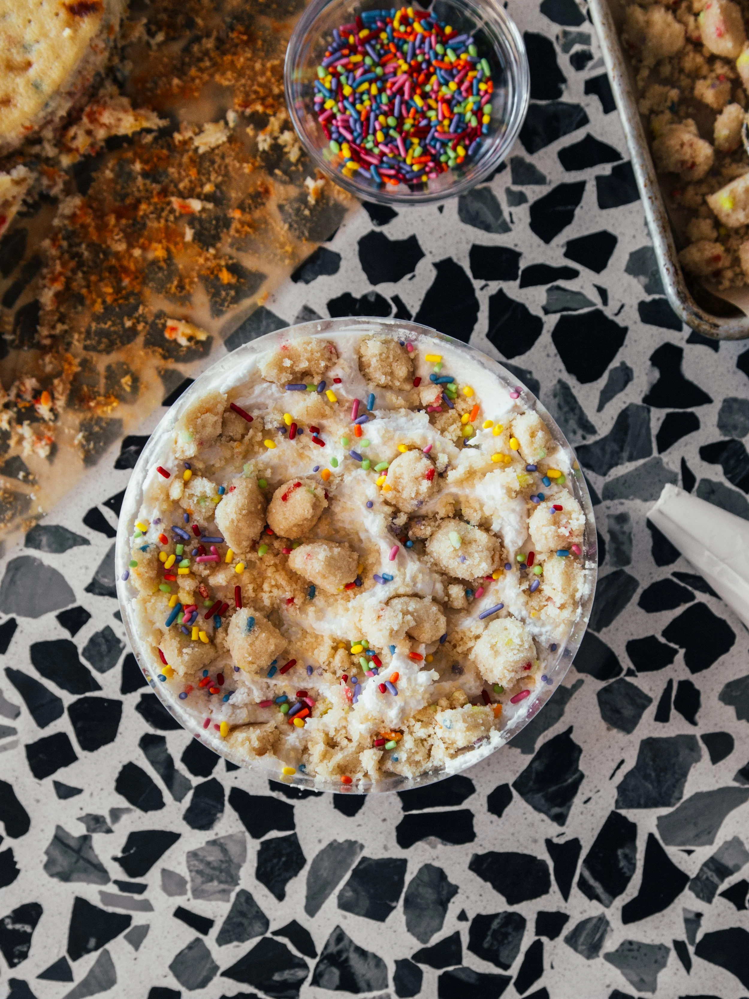 A cup of ice cream topped with colorful sprinkles on a black and white mosaic table, with additional sprinkles and baked goods around it.