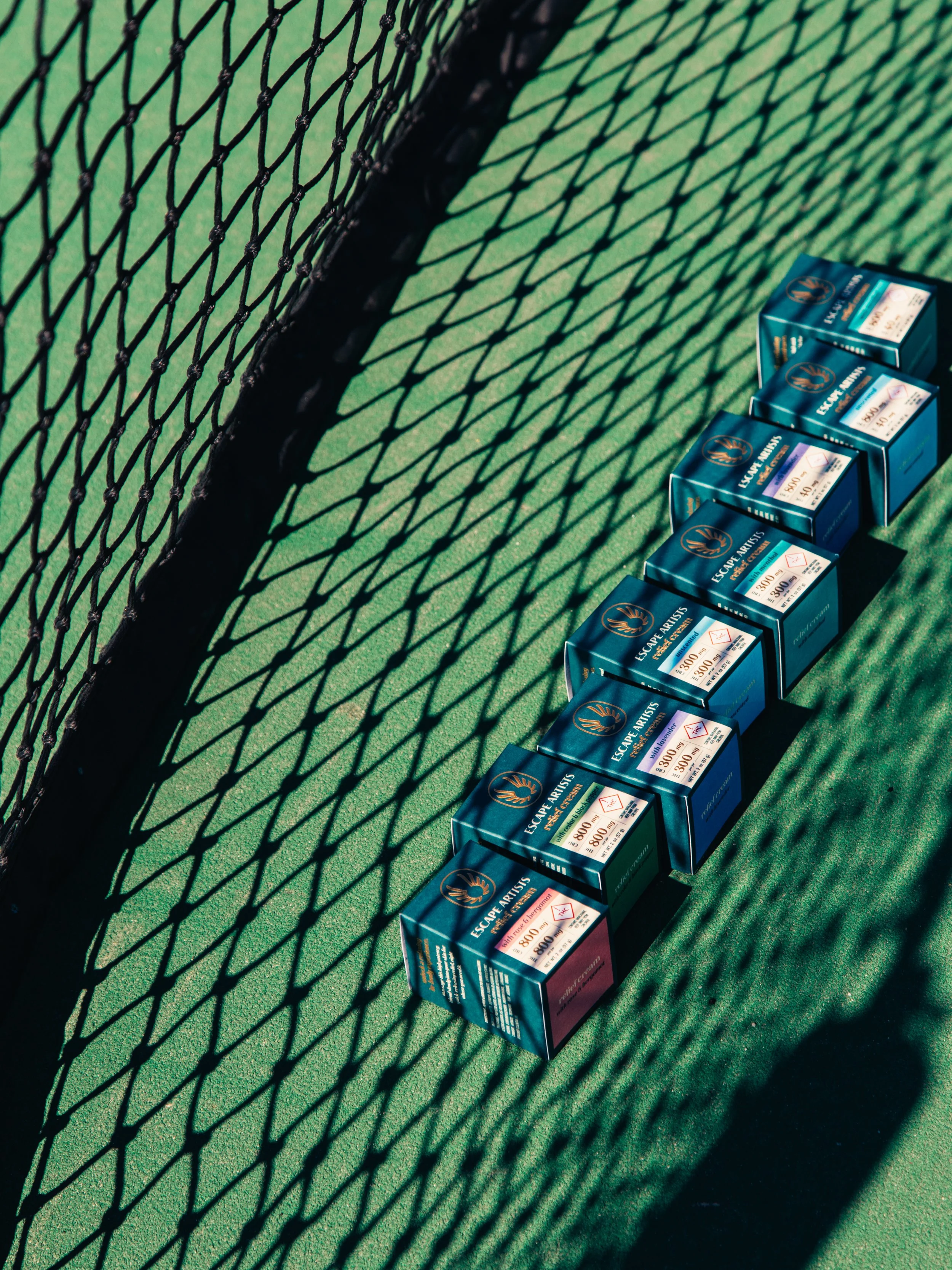 A row of infused topicals lined up on a green tennis court, with a shadow of the fence cast across the surface.