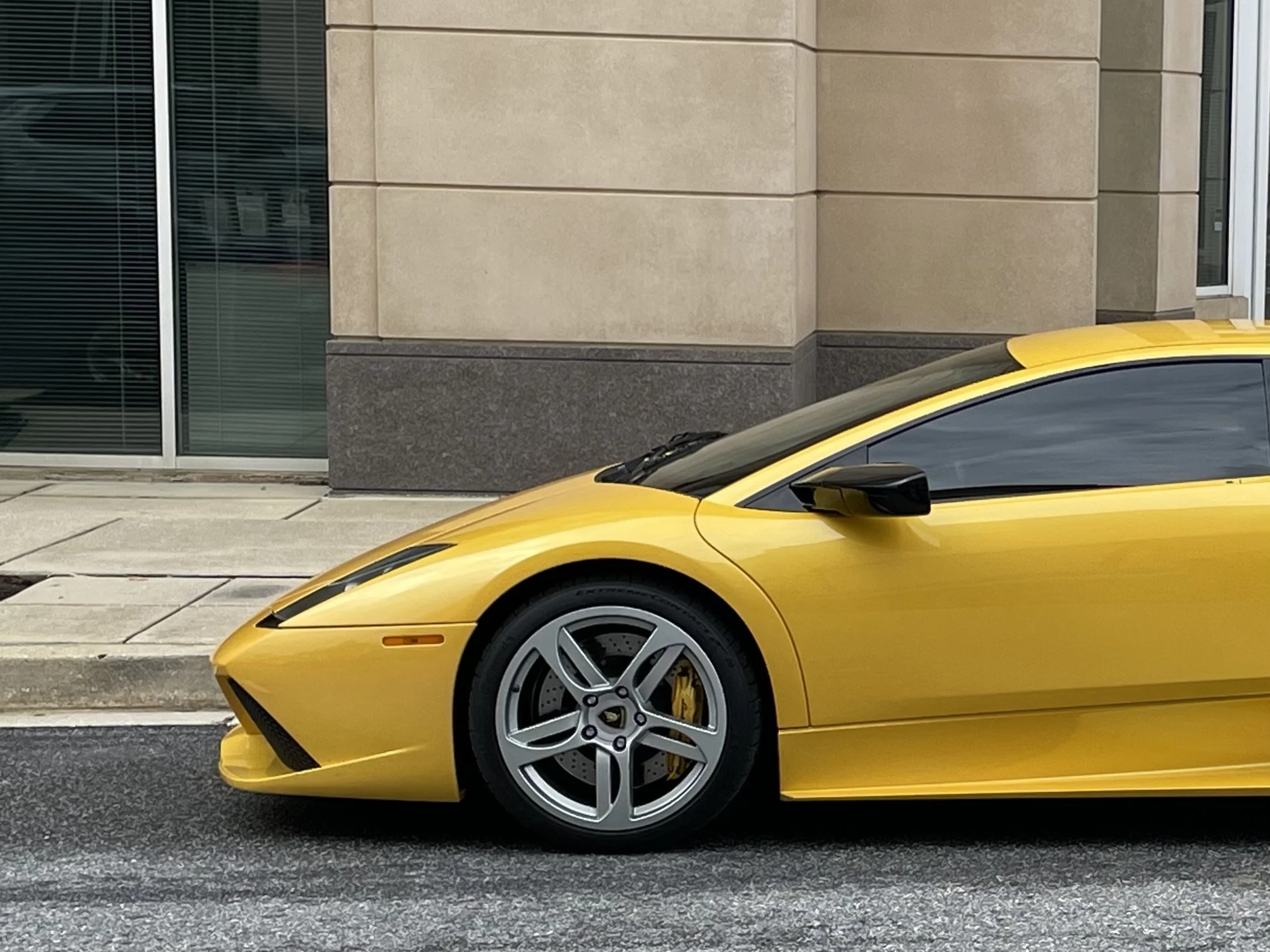 Yellow Lamborghini Murcielago car parked on city street next to a sidewalk and a modern building with large glass windows.