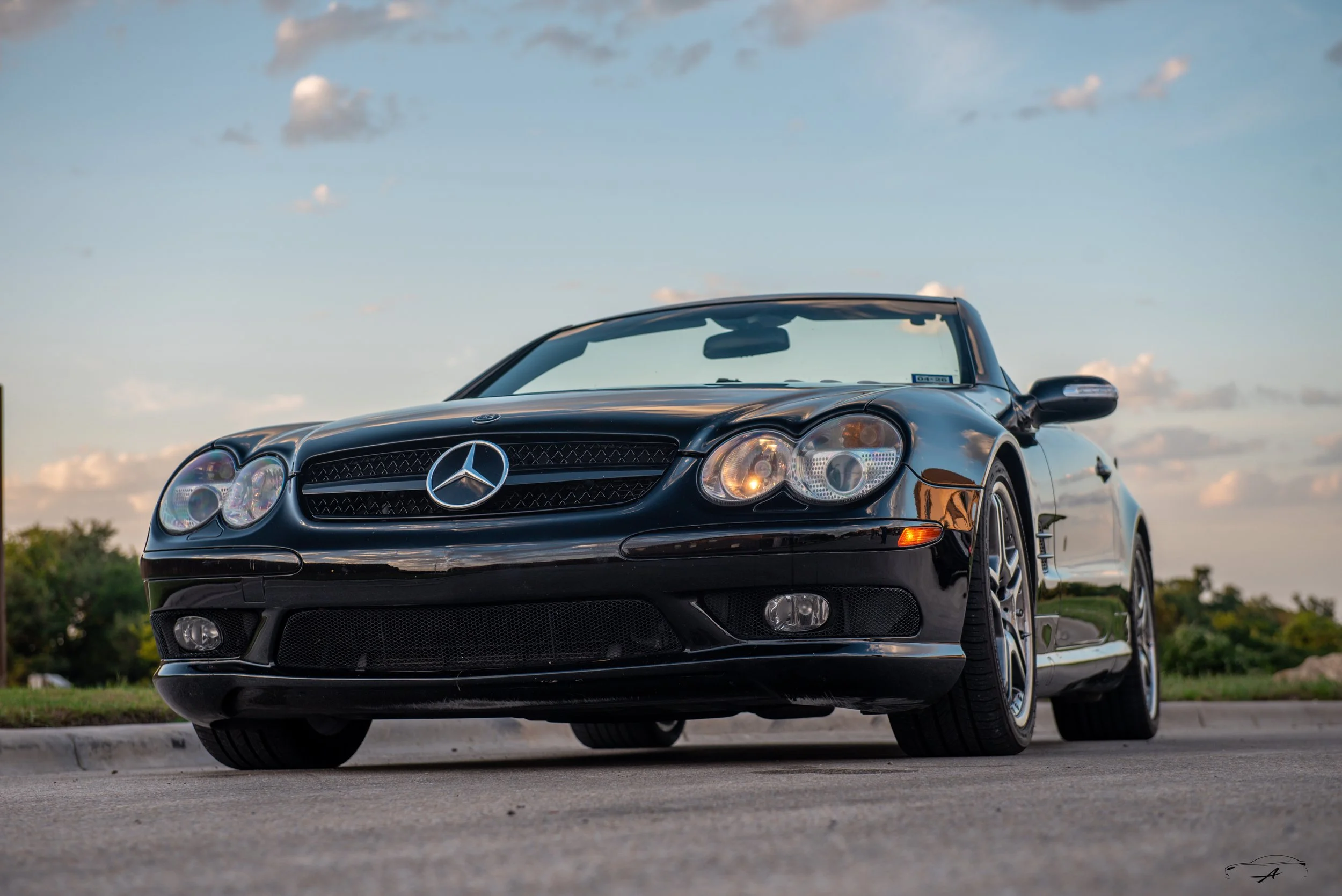 A black Mercedes-Benz convertible car parked on a road with a grassy area and trees in the background under a partly cloudy sky.