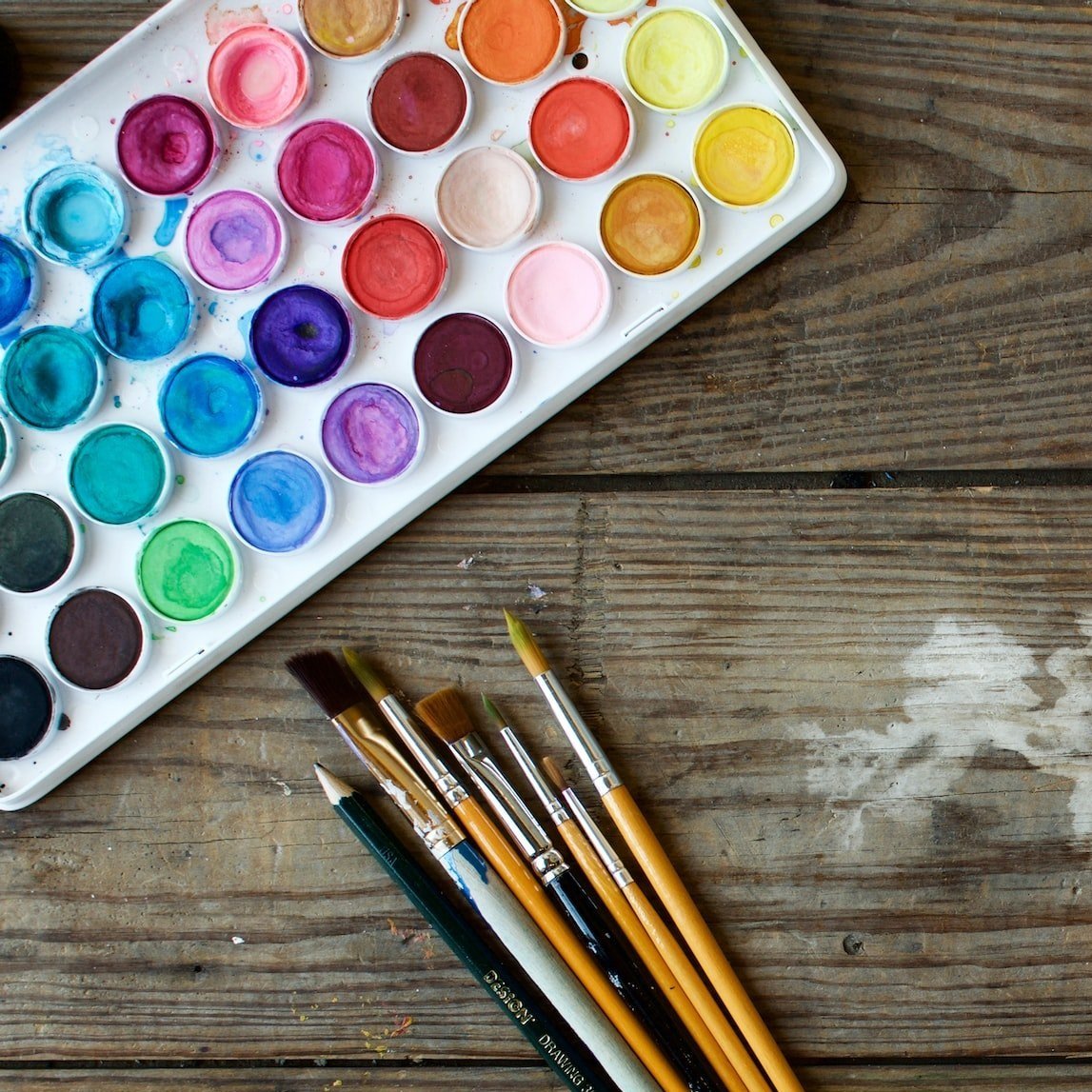 Watercolor paint palette with 24 colors and paint brushes on a wooden surface.