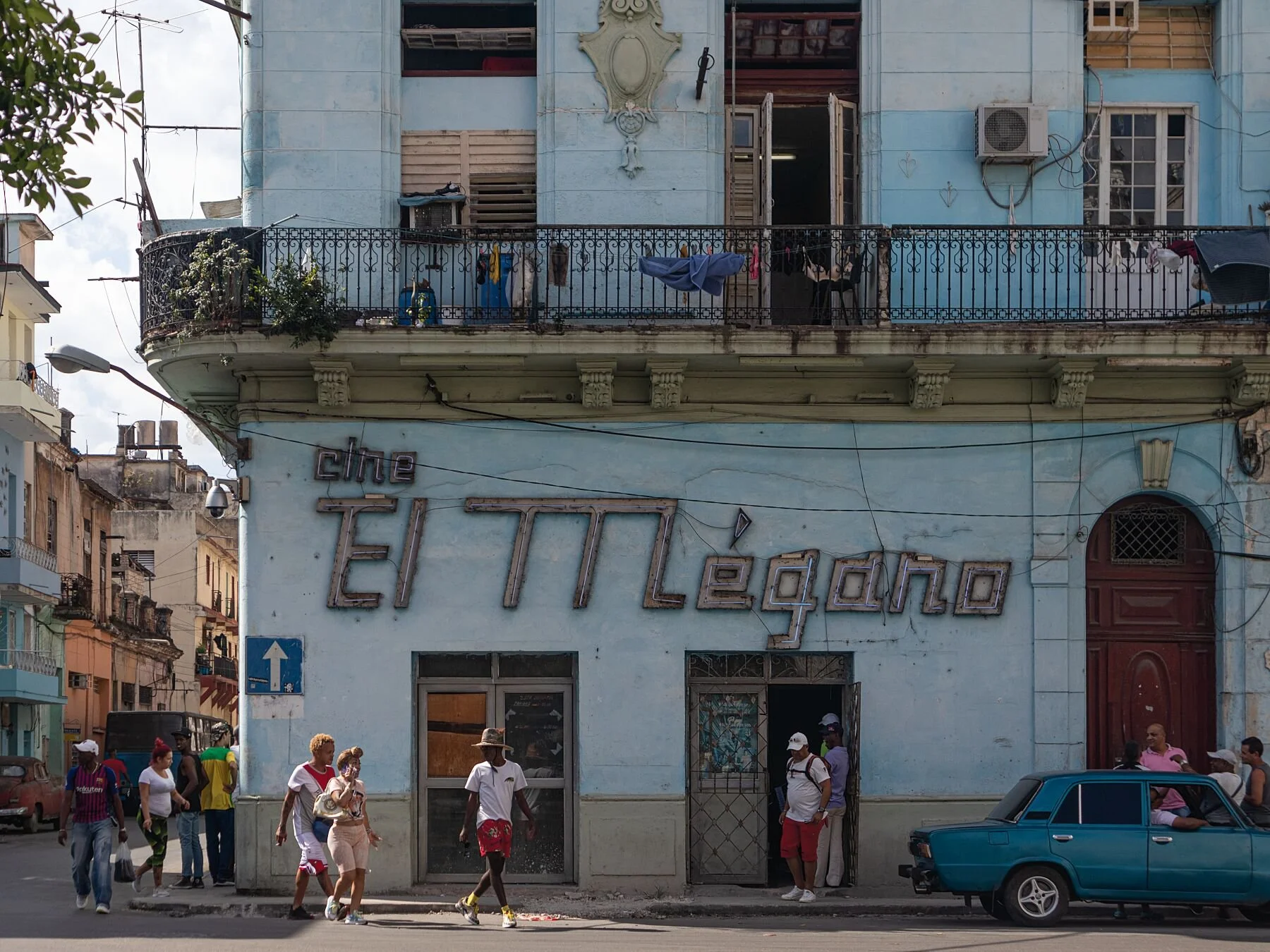 Blue, weathered building with "Cine El Mégano" sign. People walk by, and a vintage car is parked in front, conveying a nostalgic urban scene.