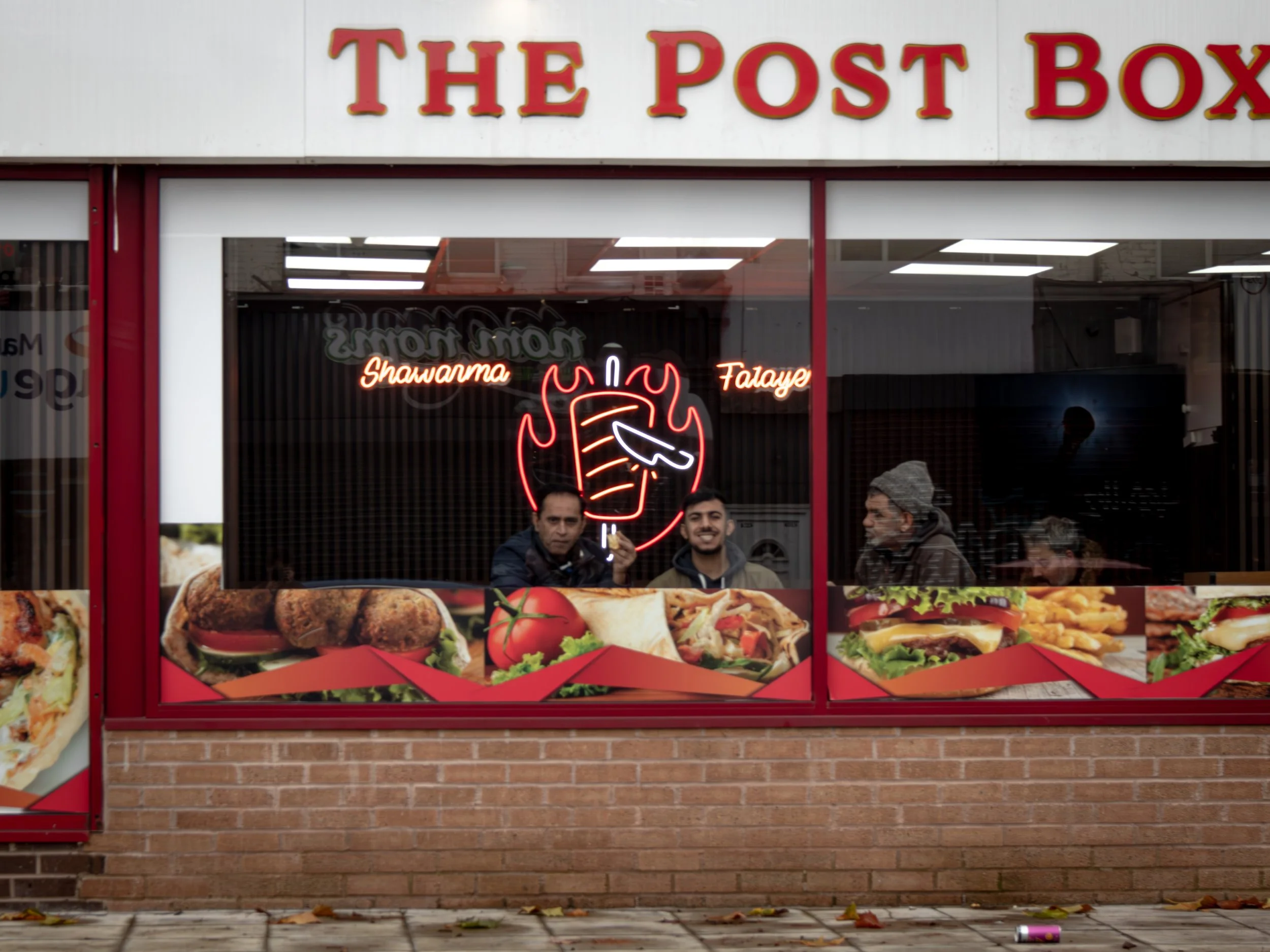 A storefront window displays a neon shawarma sign. Inside, three people sit, visible through glass adorned with food images. The mood is casual and inviting.