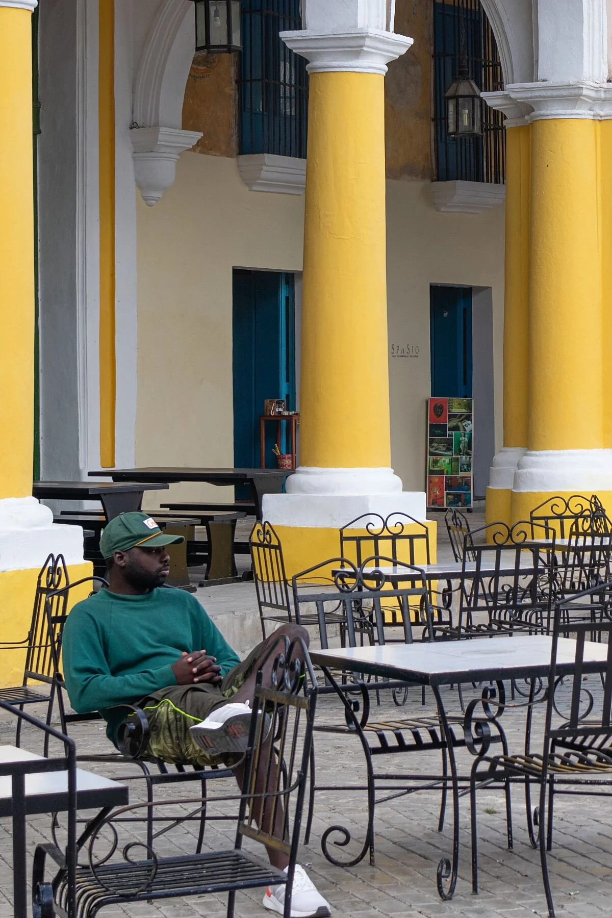 A person in a green outfit and cap sits in a wrought iron chair at an outdoor cafe with yellow columns, exuding a relaxed, peaceful vibe.