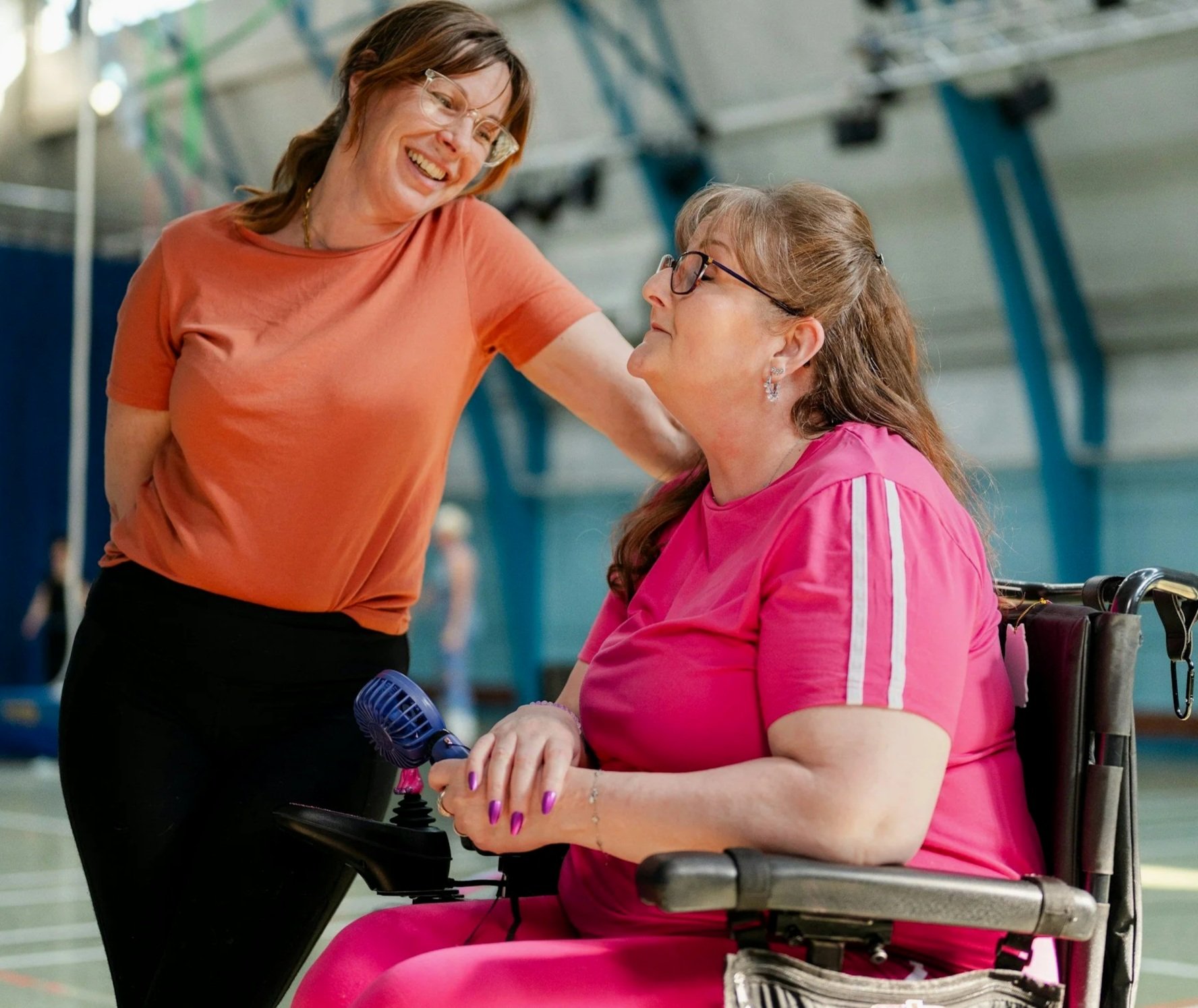 Two women in a gym: one standing and smiling, the other seated in a wheelchair, holding a joystick, with the standing woman touching her shoulder.