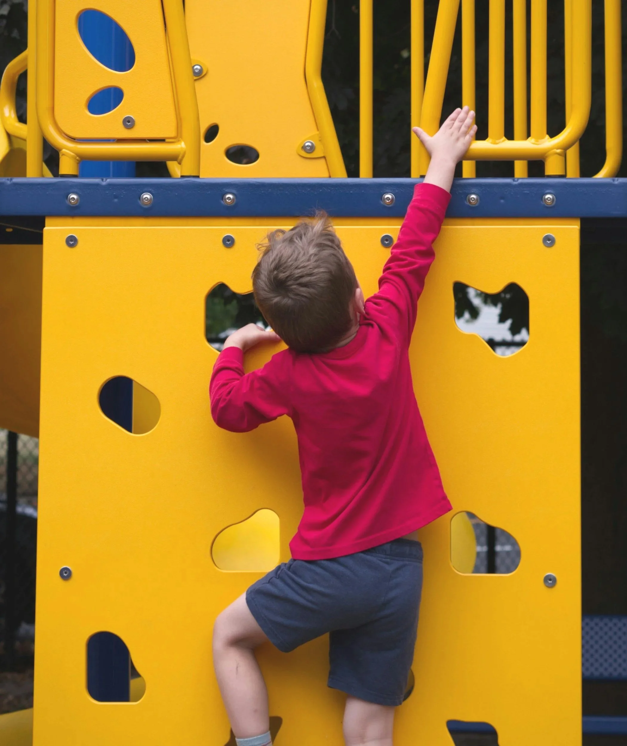 A child climbing a yellow playground structure with cut-out shapes, wearing a red long-sleeve shirt and navy shorts.
