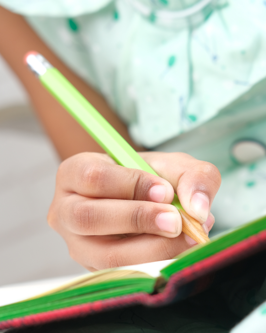 Close-up of a child's hand holding a yellow pencil and writing in a colorful notebook.