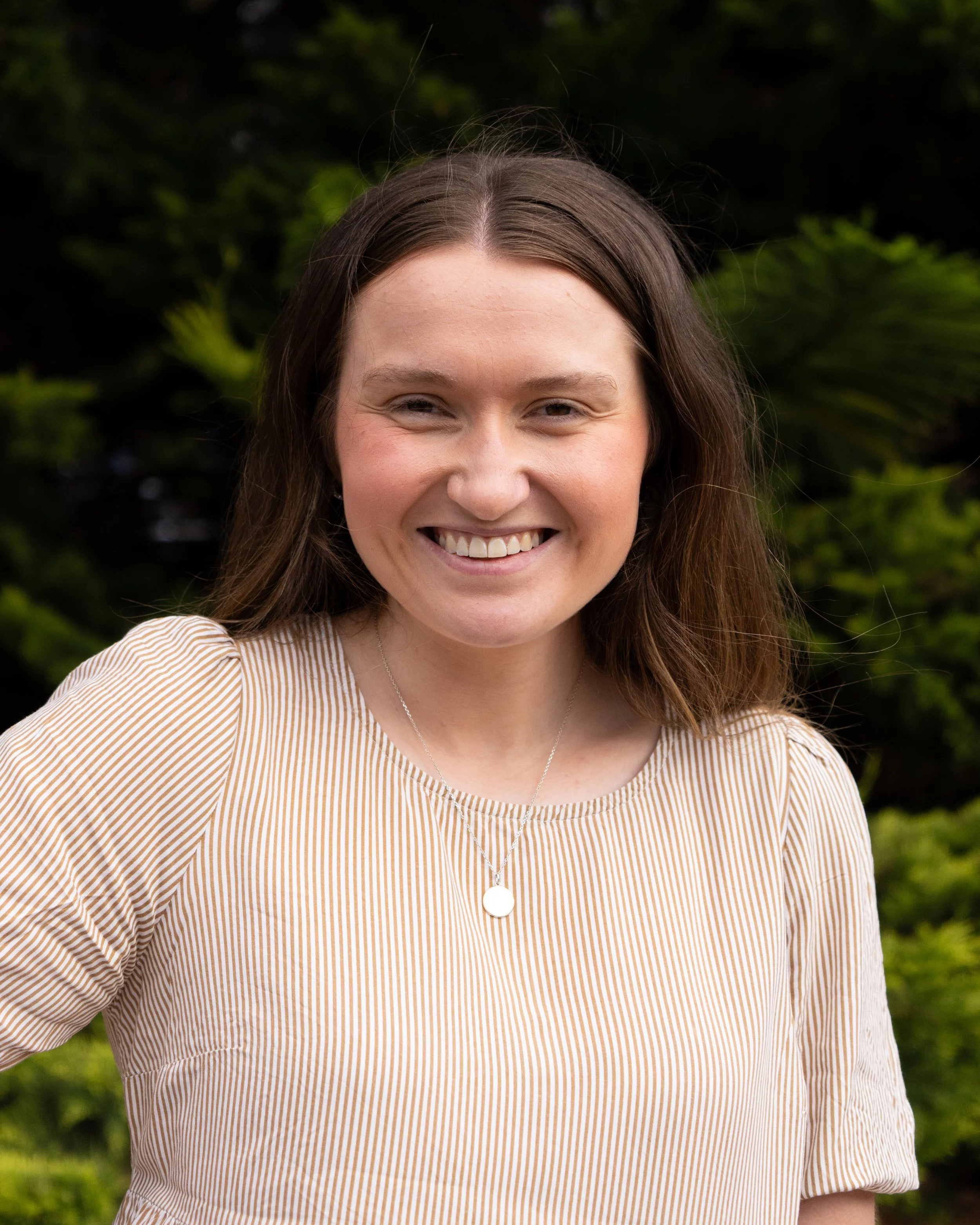 Close-up of a smiling woman with long brown hair, wearing a beige and white striped top, standing outdoors with green foliage in the background.