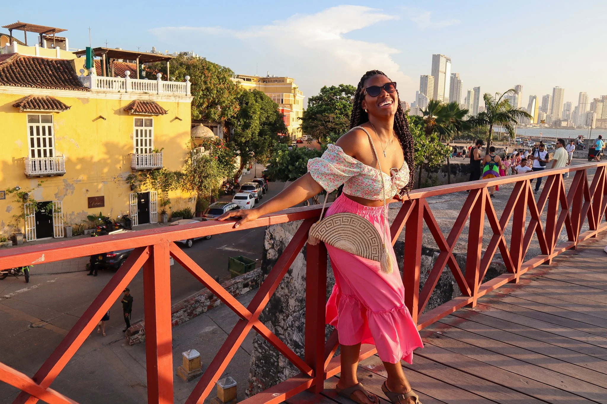 A woman smiling and posing on a wooden bridge, wearing sunglasses, a pink skirt, and a floral top, with a cityscape and a yellow building in the background.